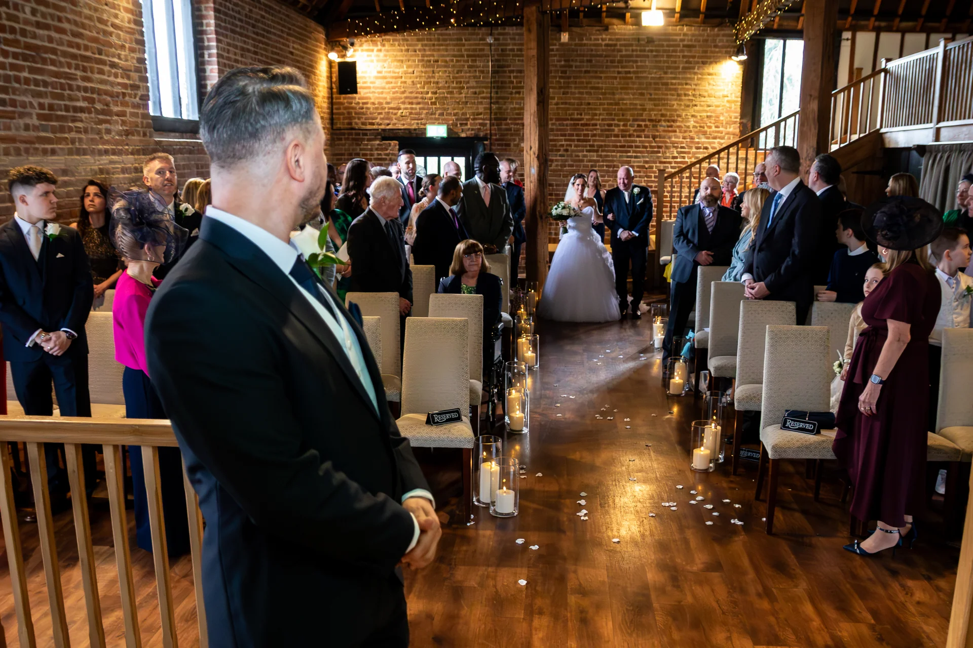 The groom looks back as the bride in a white ball gown enters down the aisle of a rustic candlelit barn wedding venue filled with seated guests.
