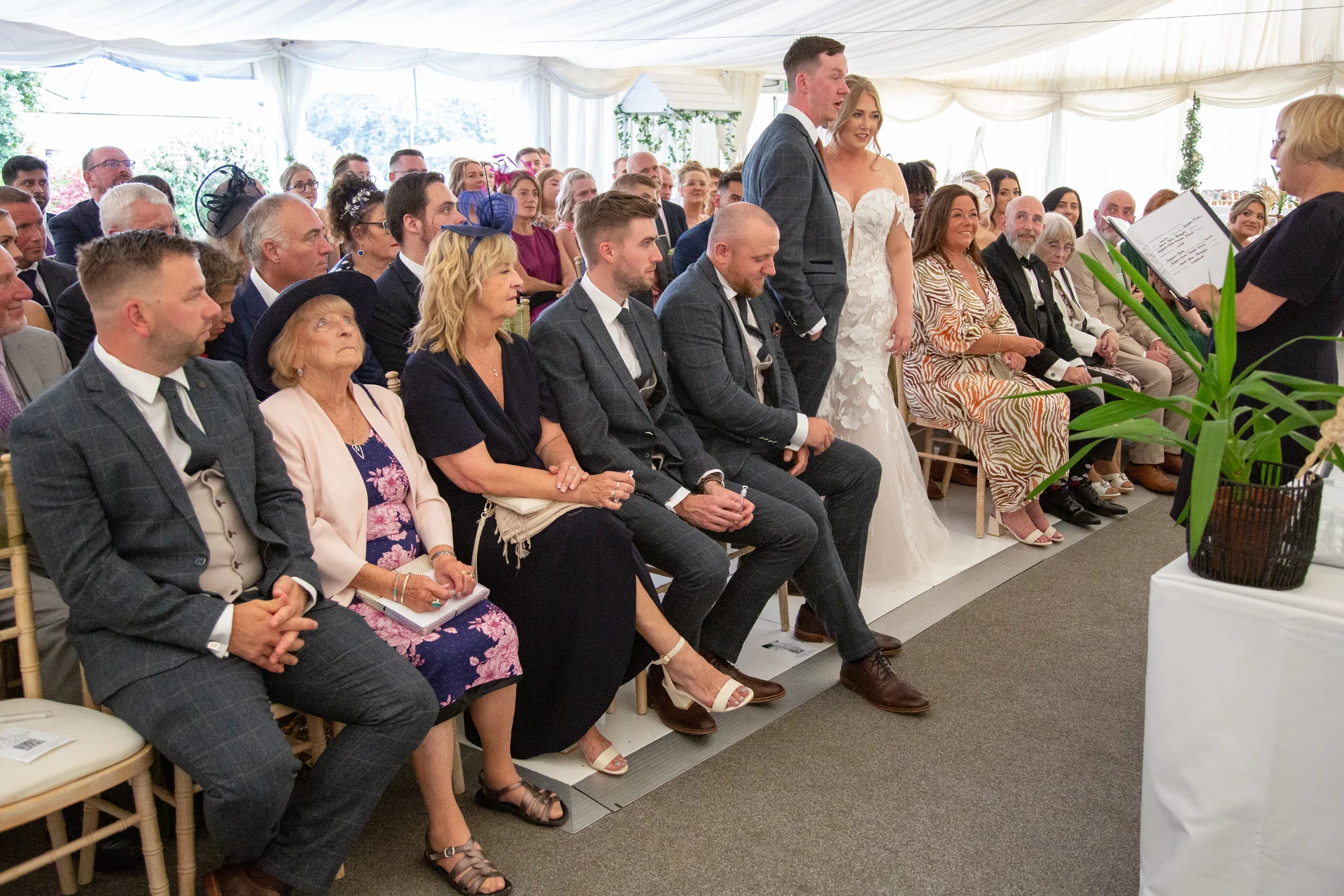 An indoor marquee wedding ceremony featuring seated guests, a couple standing near the front, and a celebrant conducting the service.