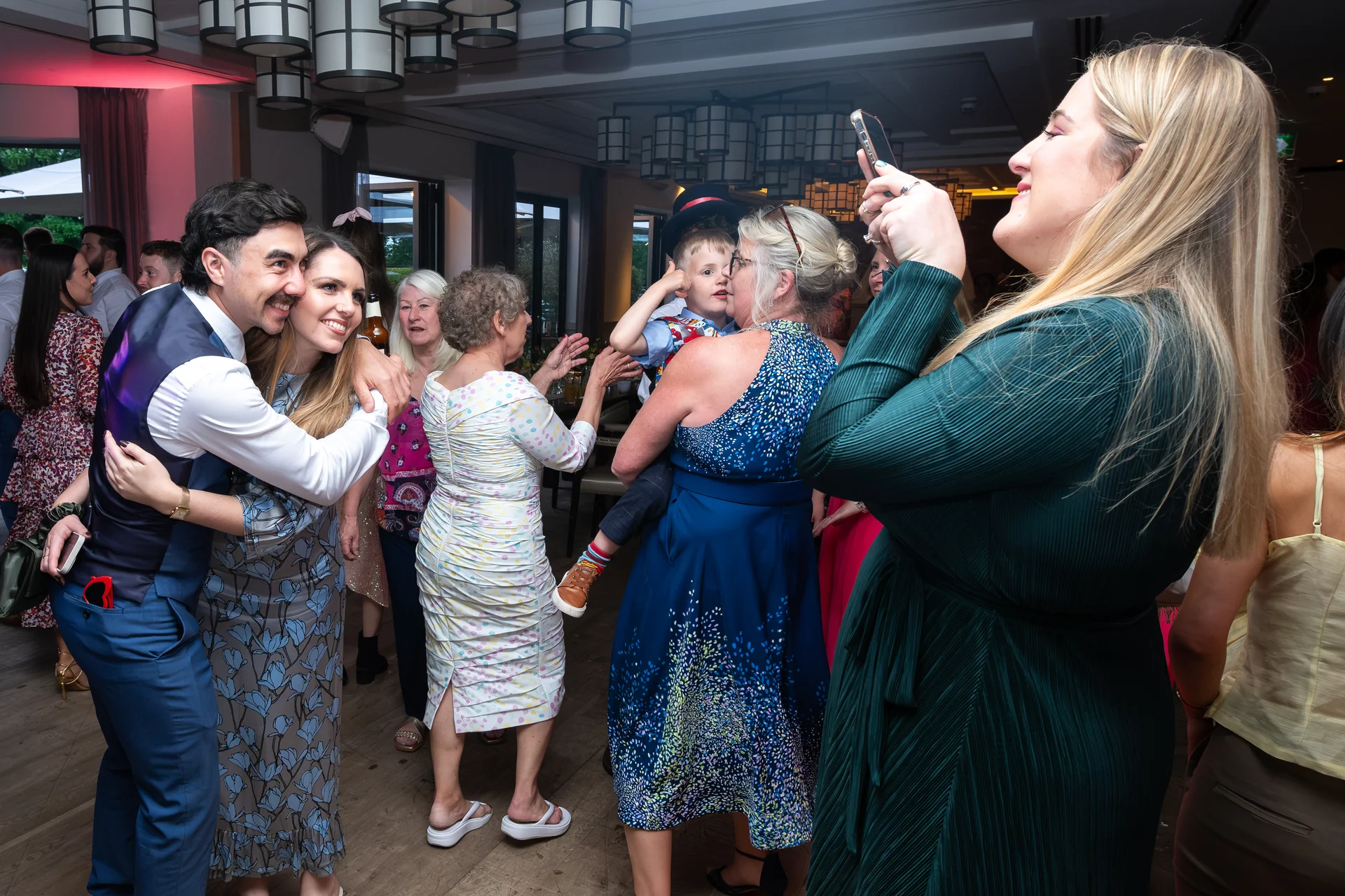 A blonde wedding guest in a dark green dress captures a joyful moment on her phone while guests mingle and embrace at an indoor evening reception.