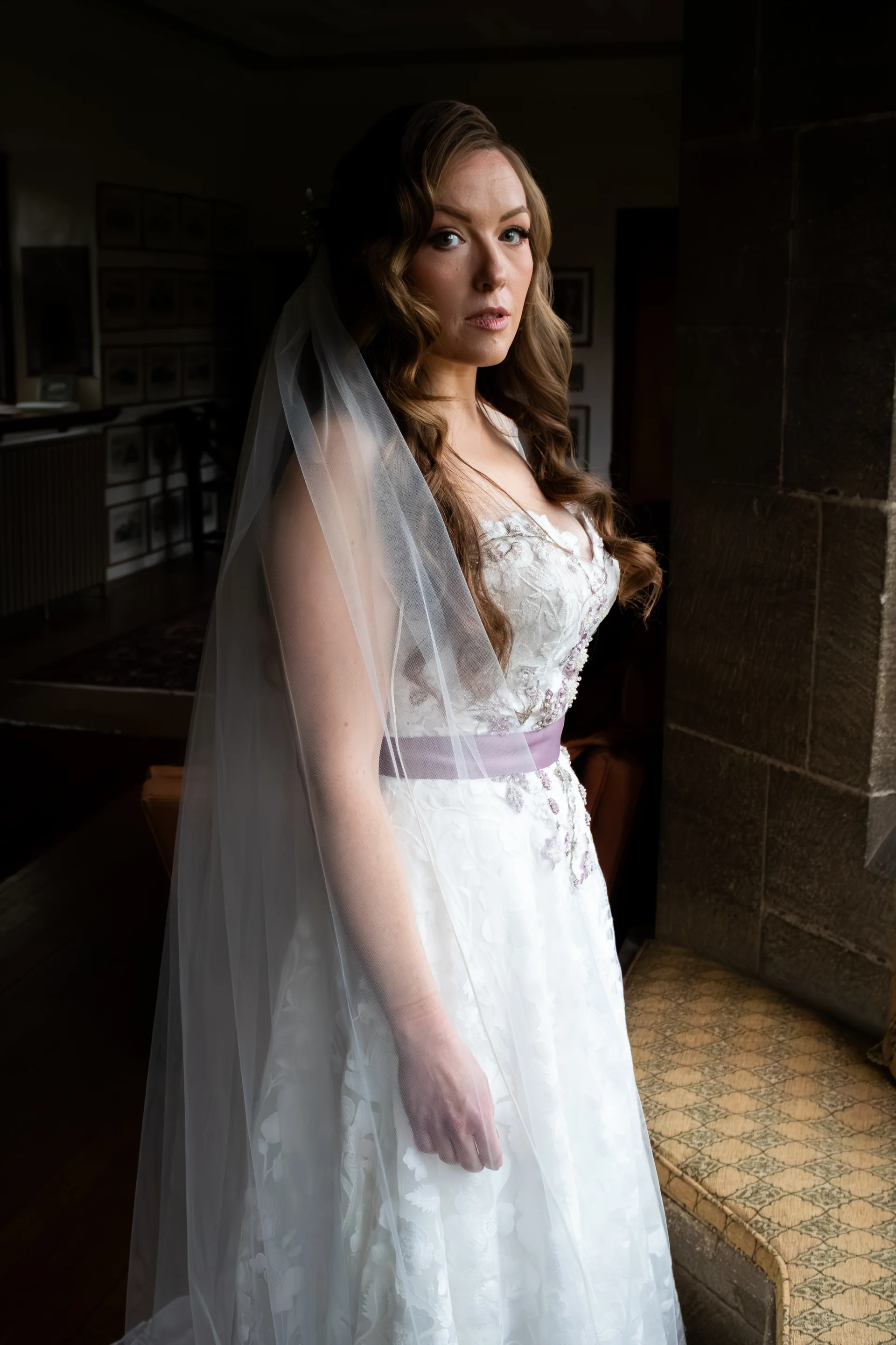 A bride stands near a stone window, illuminated by soft natural light, wearing an A-line wedding dress with floral appliqué and a long veil.
