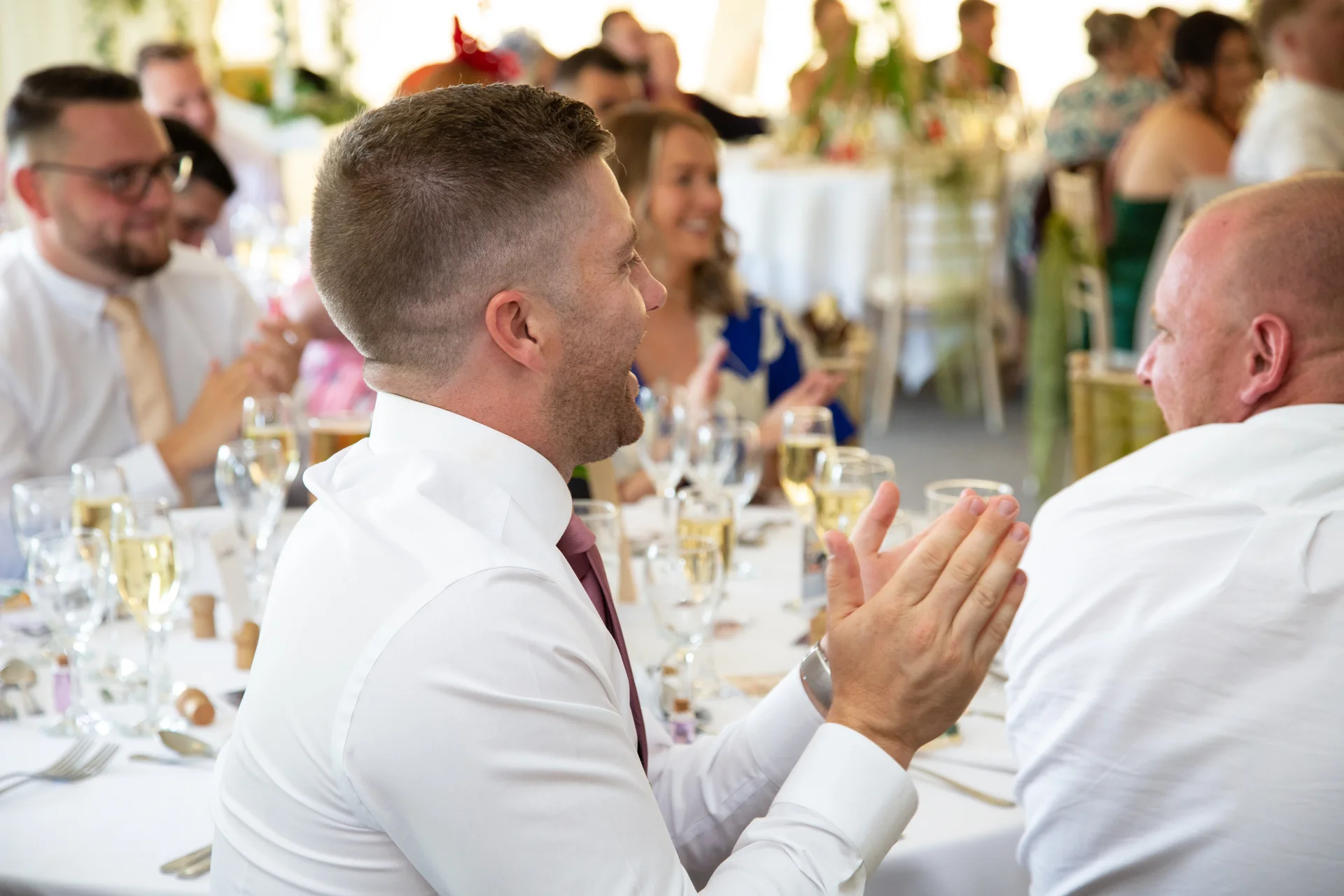 A guest in a white shirt and tie claps and laughs during the wedding breakfast speeches.