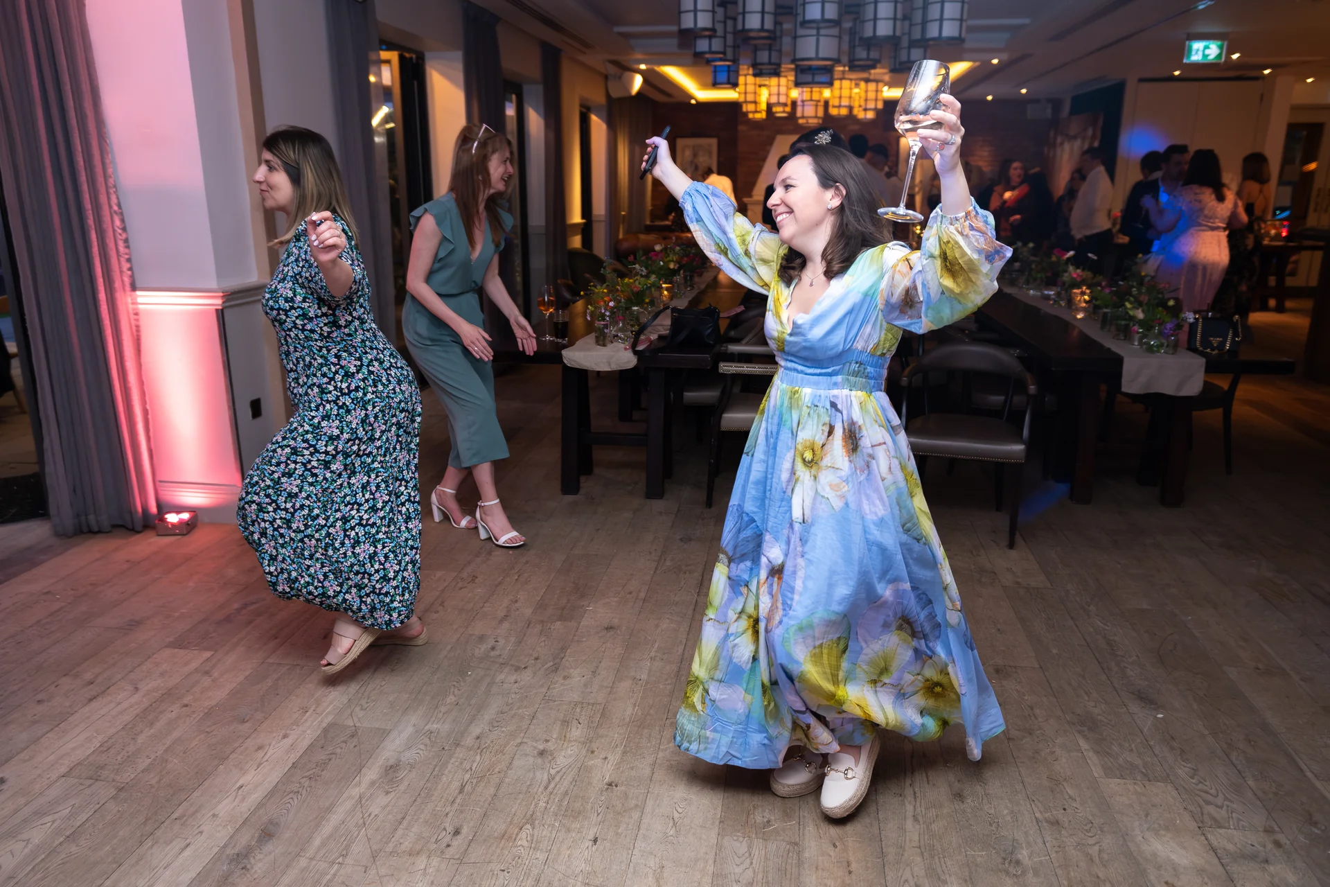 Joyful wedding guests dance enthusiastically on a wooden dance floor in a warmly lit venue, with one woman holding up a glass.