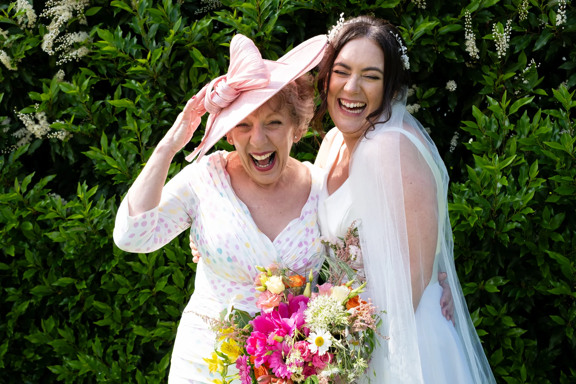 A joyful bride and a laughing mother of the bride in a pink fascinator pose together in front of lush green foliage, holding a colourful bouquet.