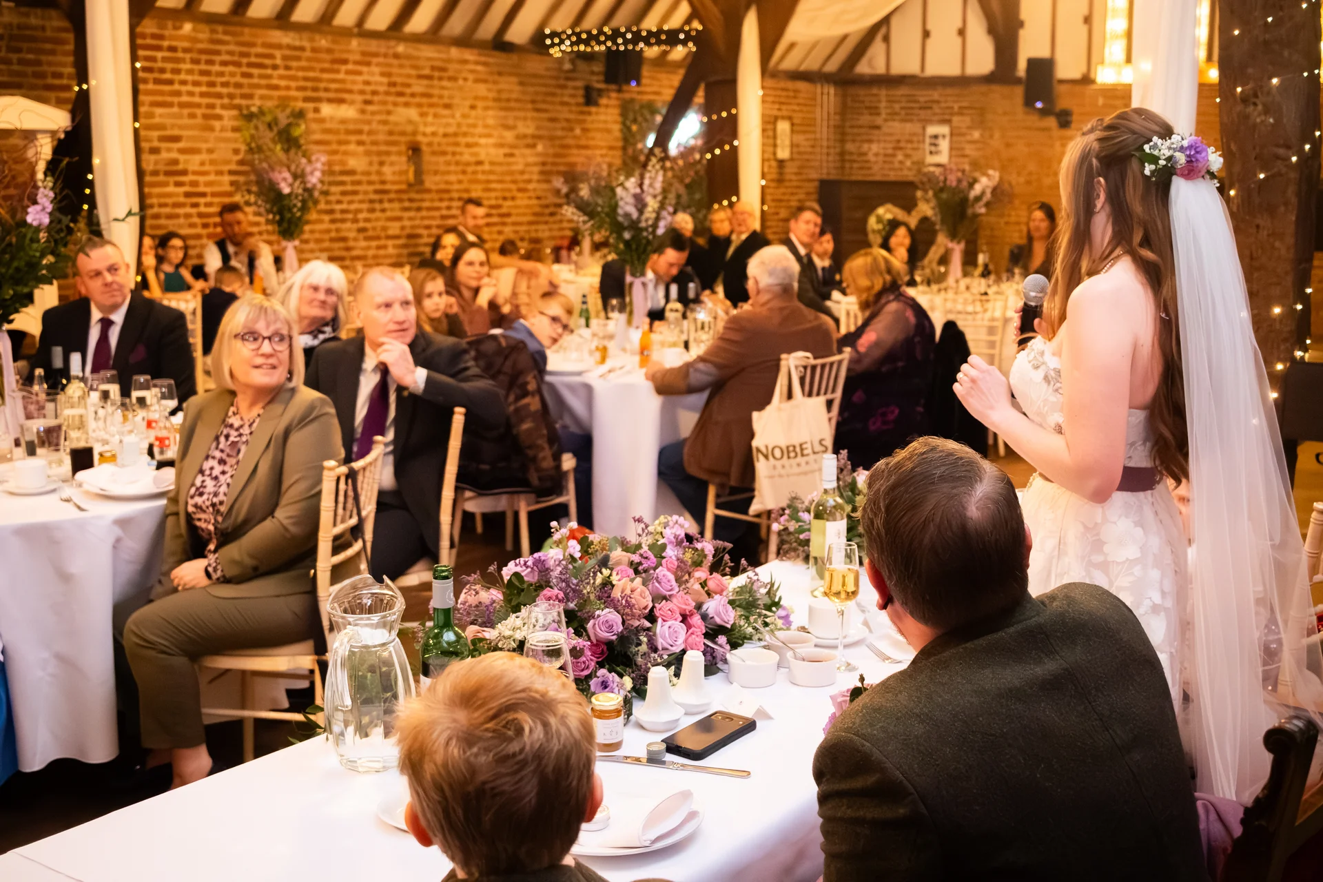 A bride gives a speech during the wedding reception in a rustic barn venue decorated with floral centrepieces and warm fairy lighting.