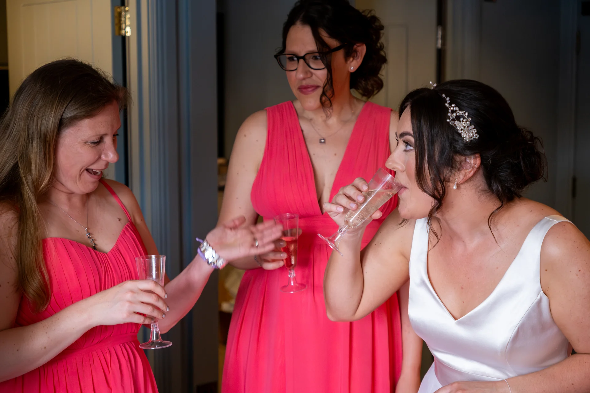 A bride in a white satin gown drinks champagne while her bridesmaids in bright pink dresses laugh and chat during morning preparations.