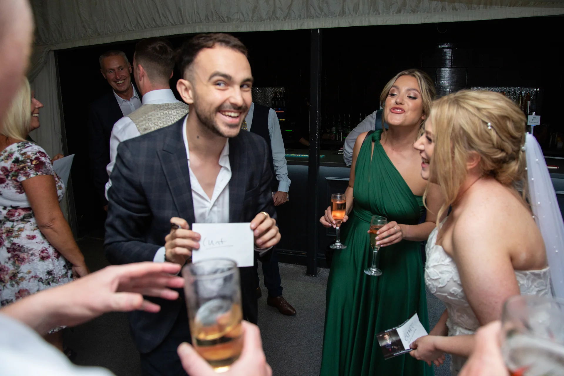 A smiling guest holds up a handwritten note while mingling with the bride and bridesmaids during the evening wedding reception.