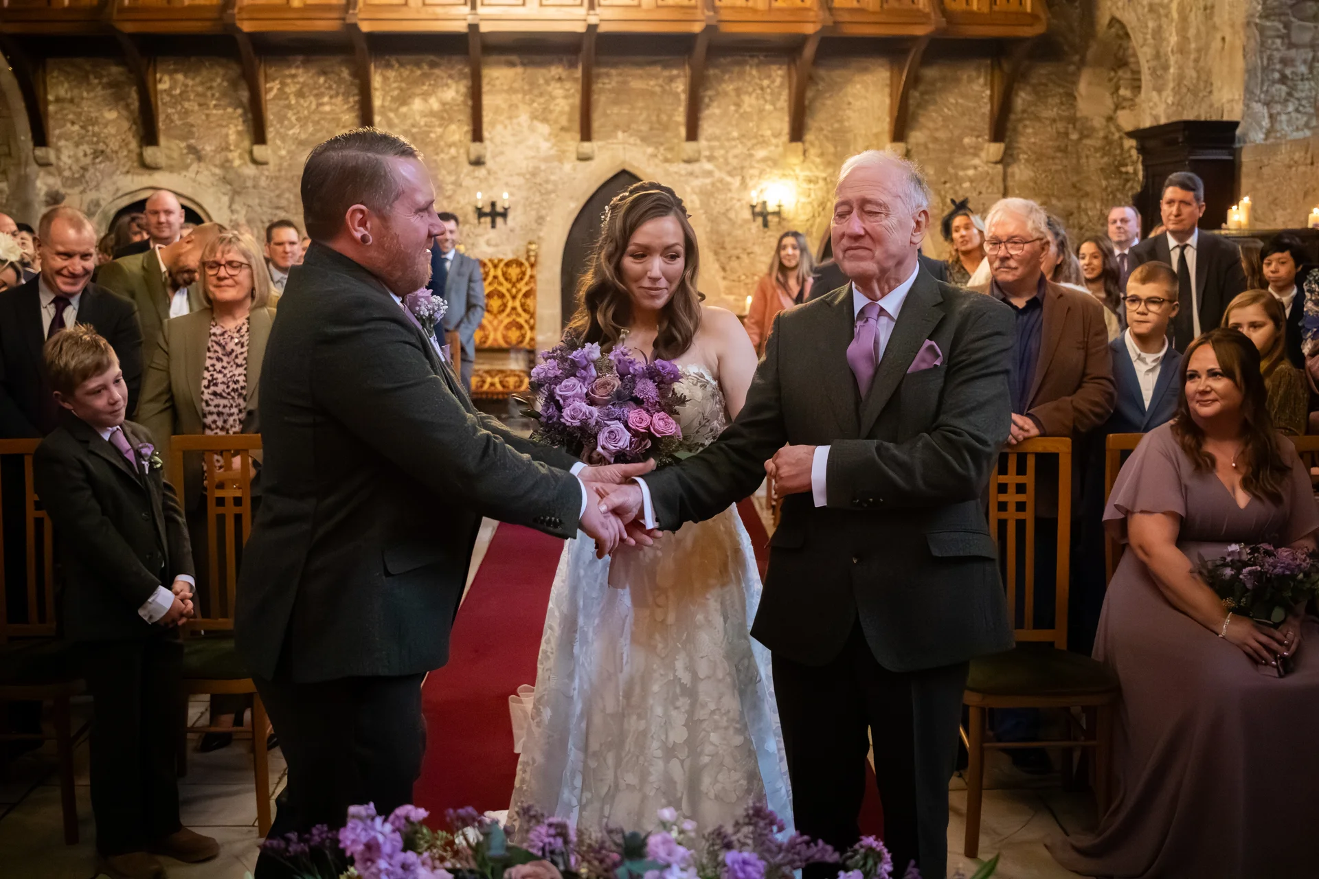 The groom warmly shakes hands with the emotional father of the bride at the altar while the bride looks on holding a purple bouquet inside a historic castle venue.
