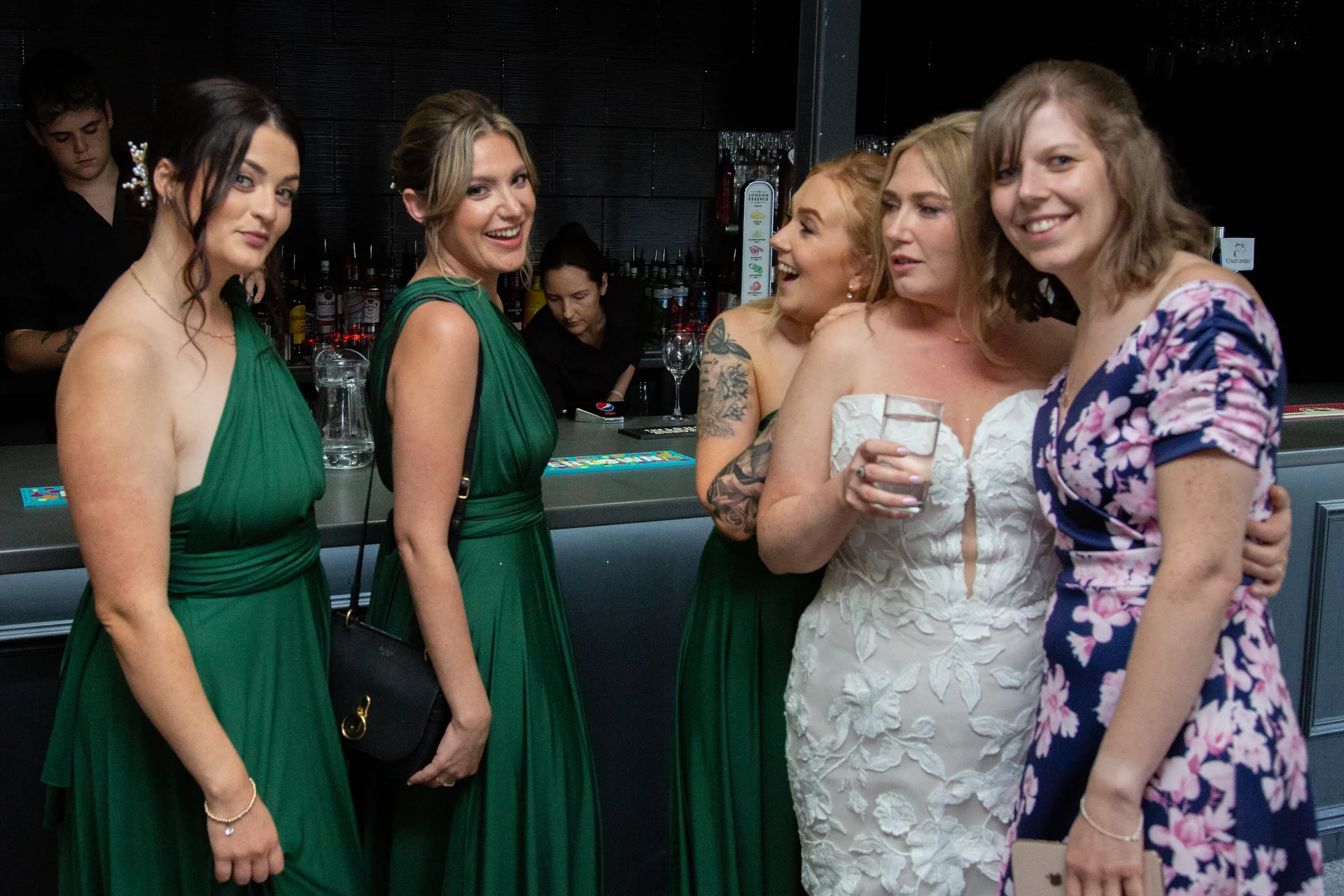 A group of bridesmaids in green dresses and a guest in a floral dress gather around a bride at the wedding bar.