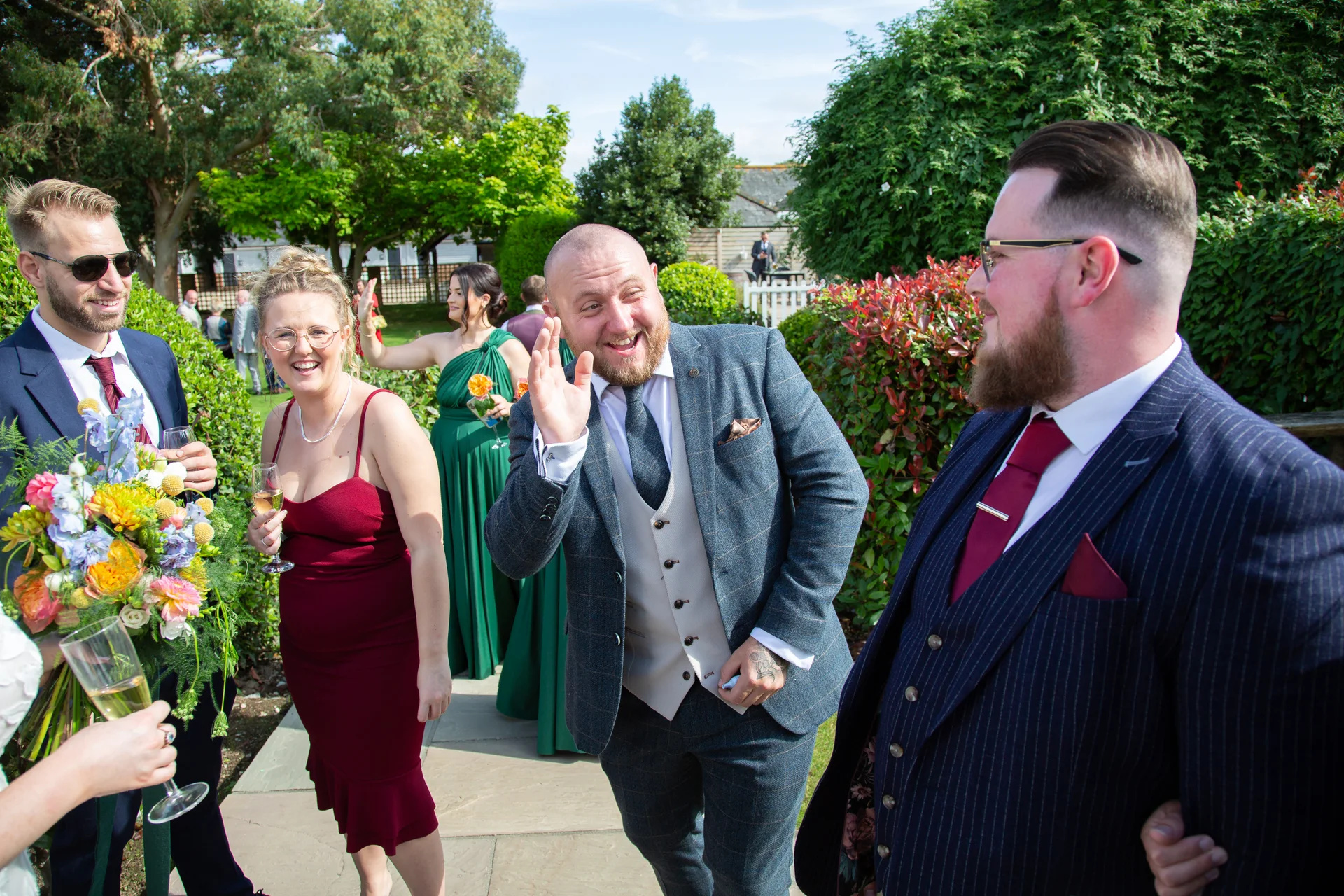 A joyful group of wedding guests and groomsmen in tweed and pinstripe suits laughing and chatting during an outdoor drinks reception in a sunny garden setting.
