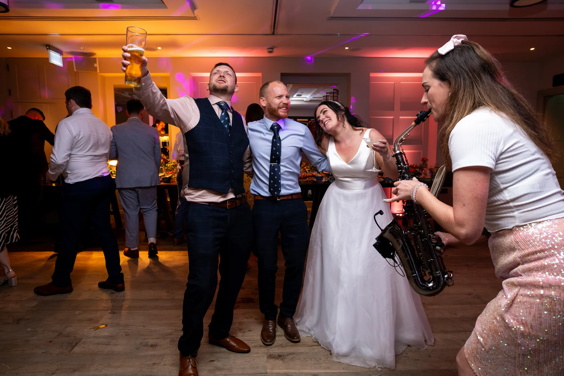 A lively wedding reception dance floor scene where guests and a bride in a white gown celebrate alongside a live saxophonist under colourful disco lights.