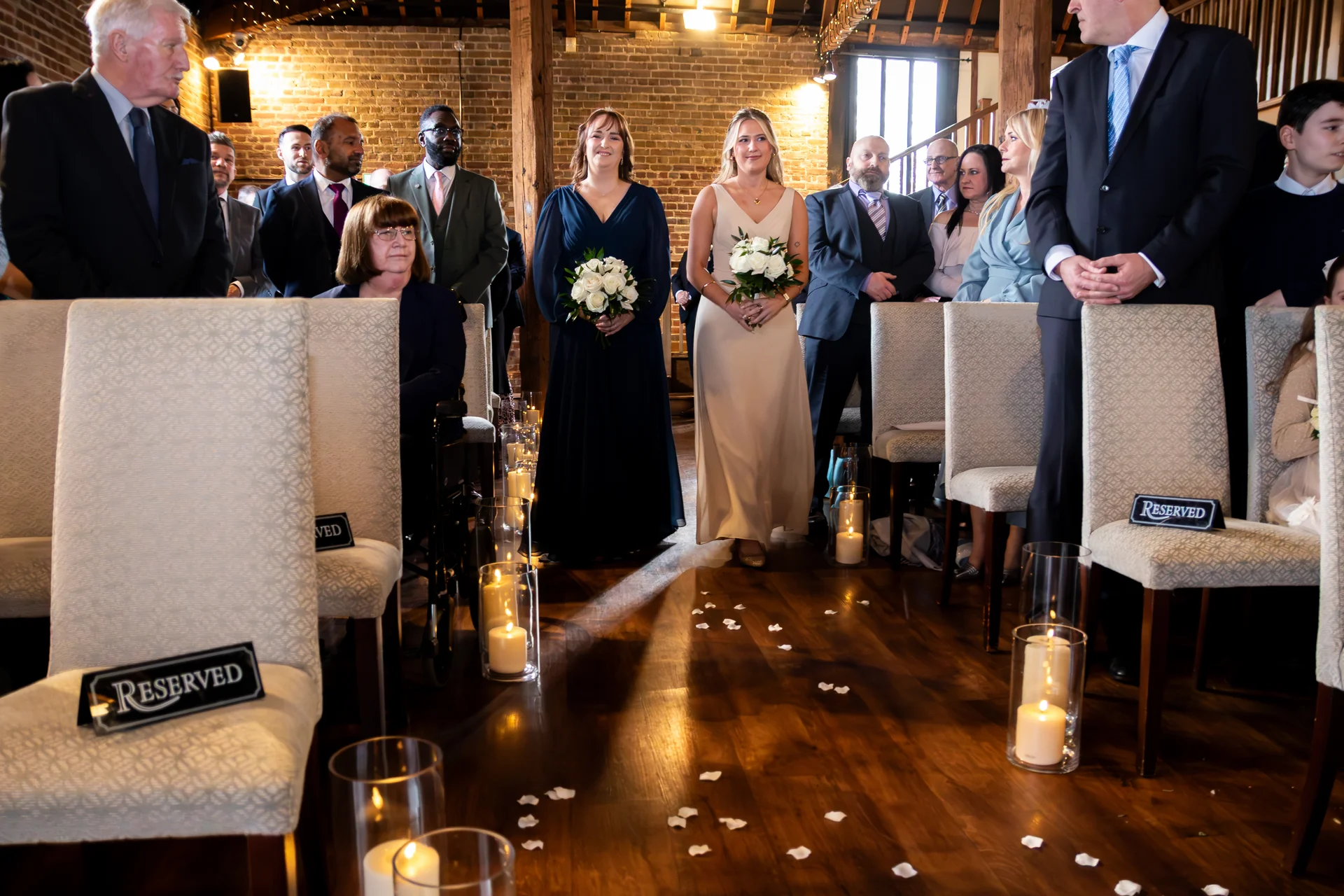 Bridesmaids holding bouquets walk down a candlelit wooden aisle inside a rustic barn wedding venue.