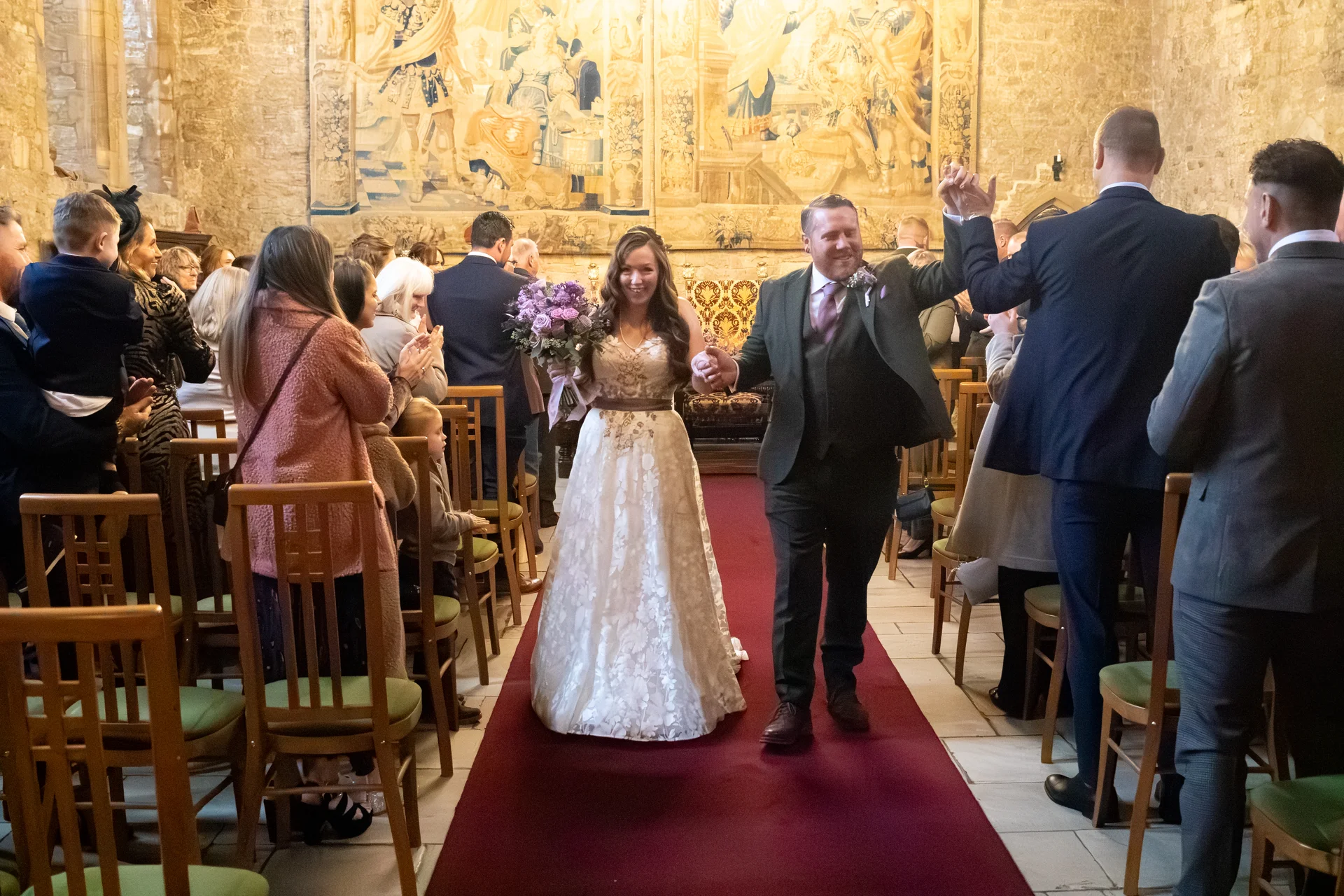 A newlywed couple joyfully walks hand in hand down the red-carpeted aisle of a historic castle venue while cheering guests applaud.