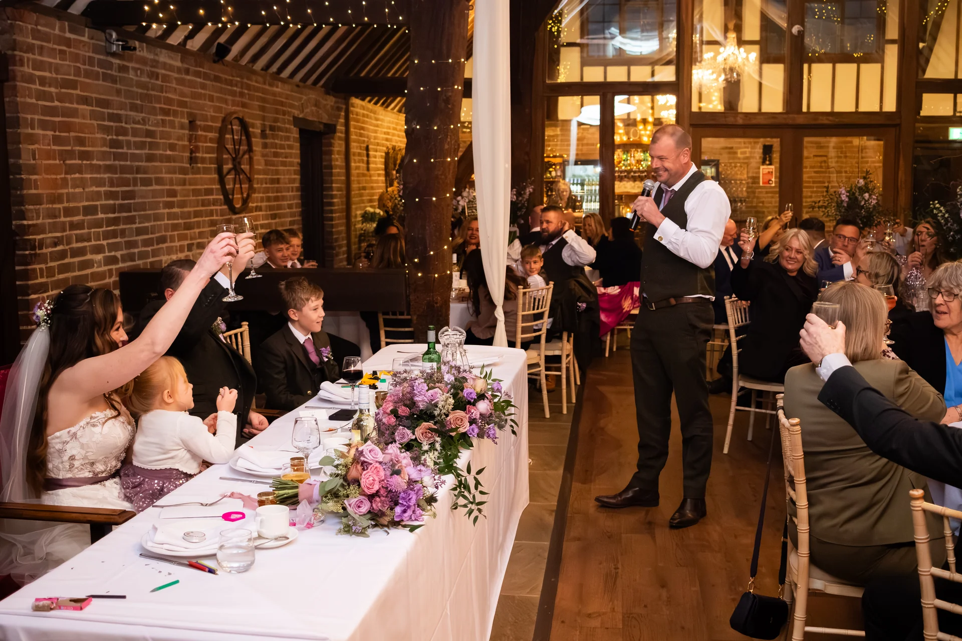 A groom delivers a speech holding a microphone during the wedding breakfast inside a rustic barn decorated with fairy lights and floral table runners.