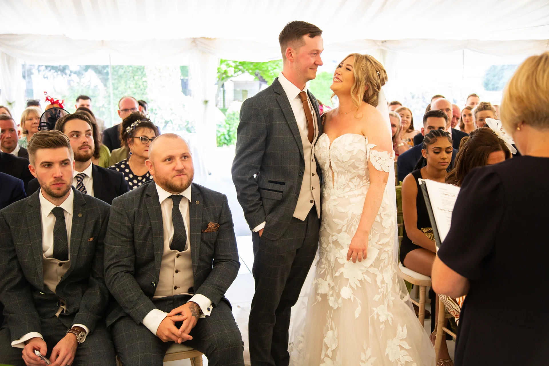 A smiling bride and groom stand together during a marquee wedding ceremony, surrounded by seated groomsmen in matching checked suits.