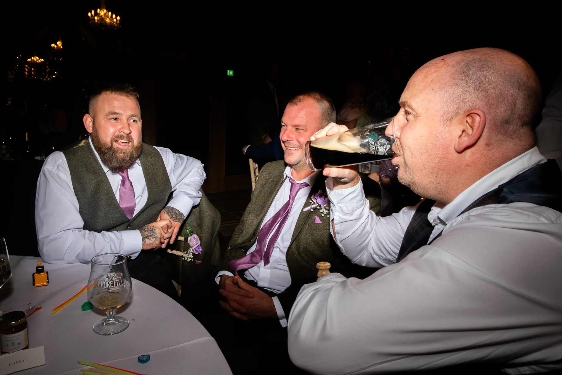 Three male wedding guests wearing tweed waistcoats and purple ties share a cheerful moment around a banquet table while one drinks a pint of dark beer.