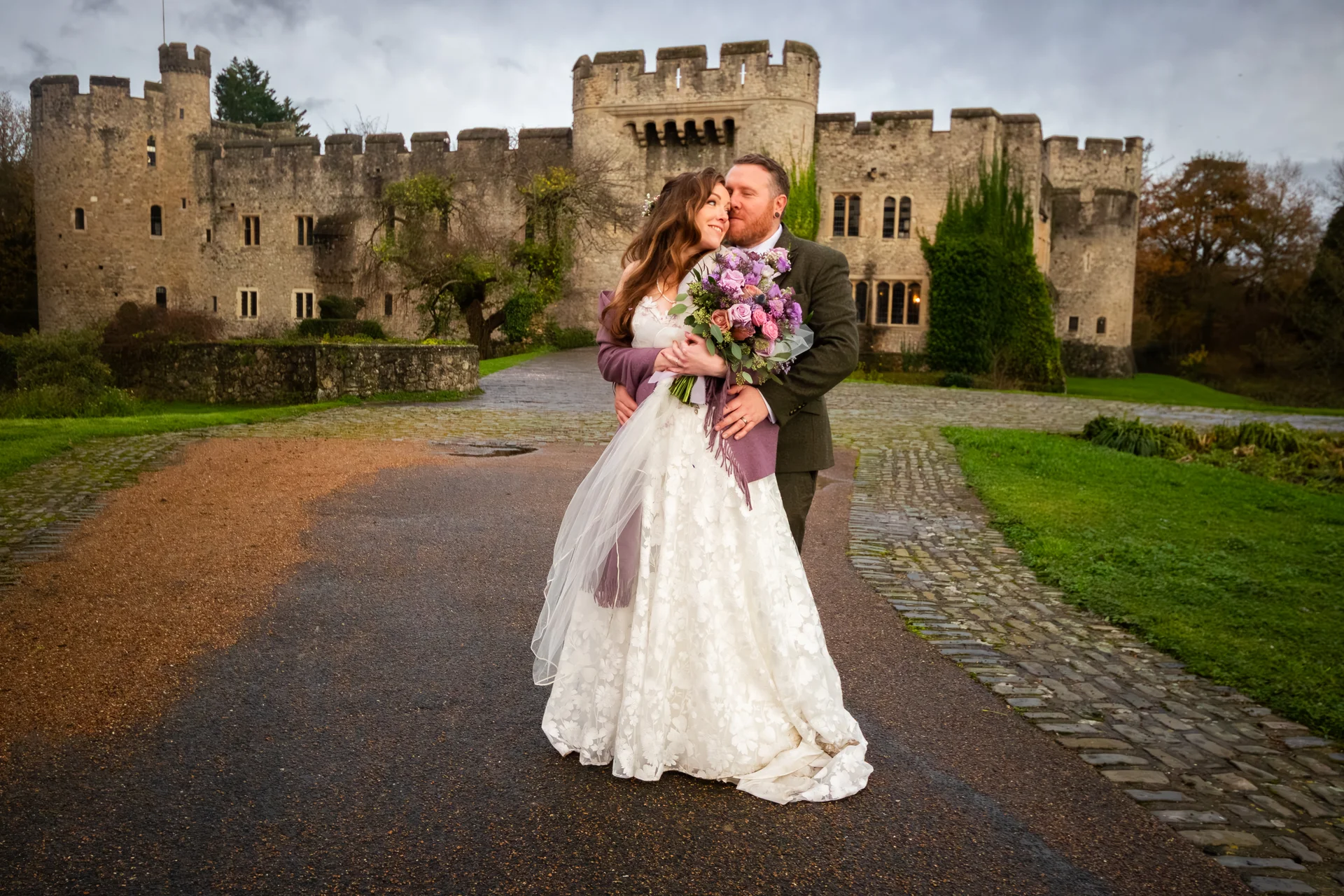 A newlywed couple embrace on a paved driveway in front of a grand historic stone castle, with the groom kissing the bride's forehead while she holds a colourful bouquet.