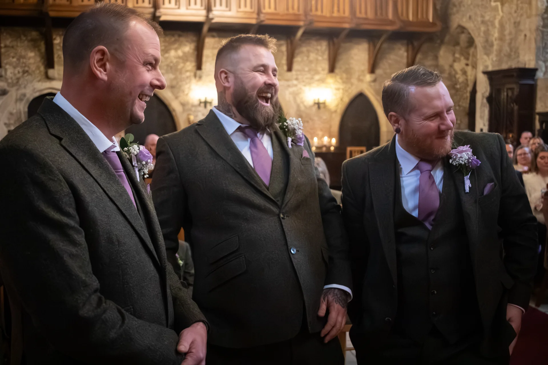 Three groomsmen in matching olive tweed three-piece suits and purple ties share a joyful belly laugh together inside a historic stone venue.