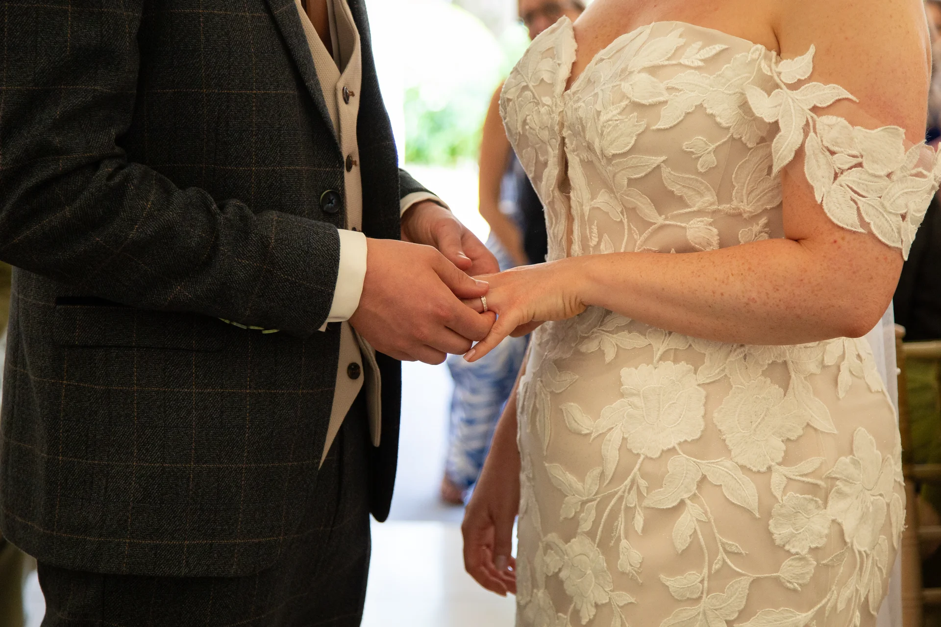 Close-up shot of a groom sliding a wedding ring onto the bride's finger during the ceremony, with the bride wearing a strapless gown featuring 3d floral lace and the groom in a checked suit and tweed waistcoat.