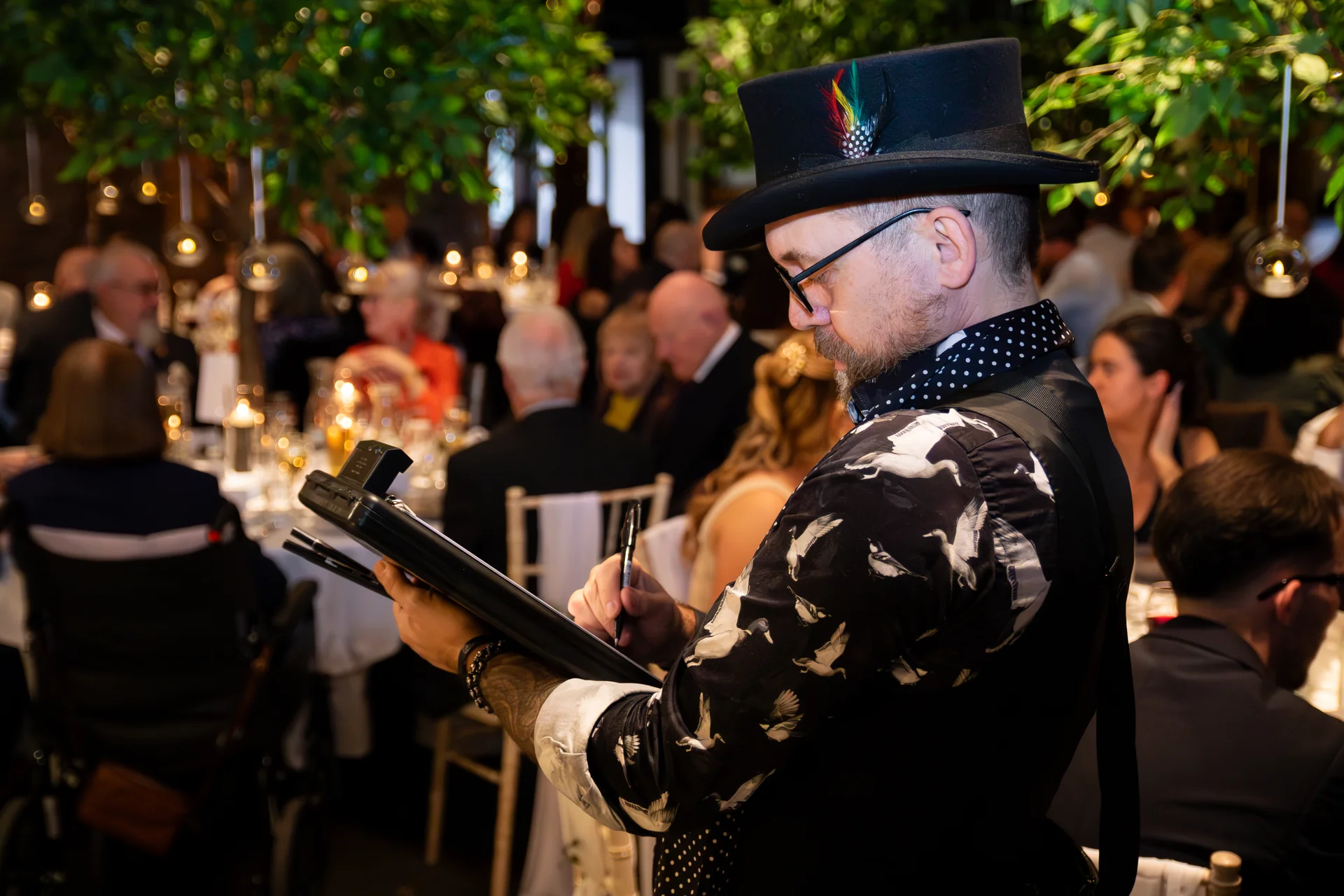An artist wearing a top hat and patterned shirt sketches at a wedding reception table filled with candlelight and guests.