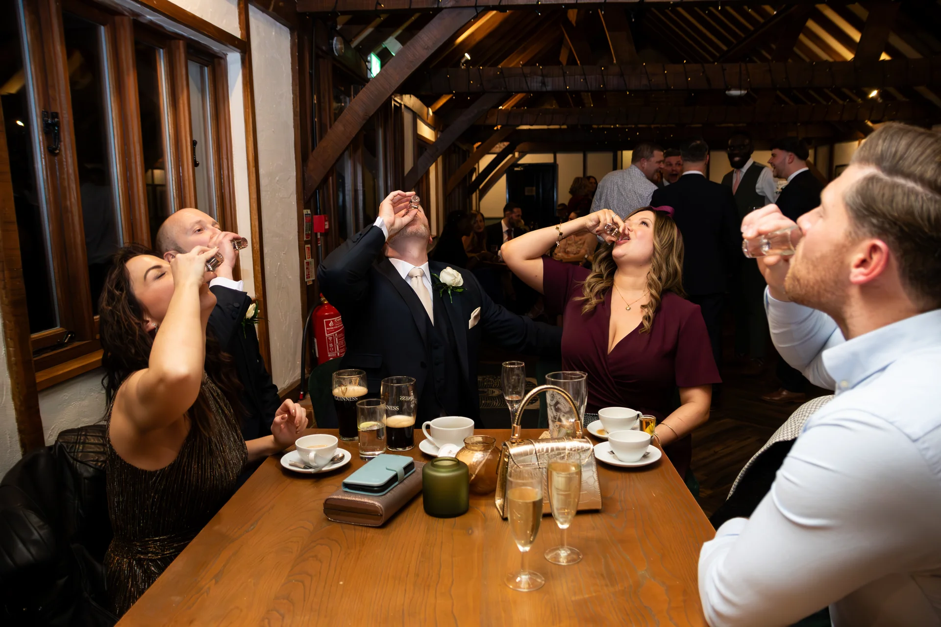 A lively group of wedding guests tilt their heads back to take shots together around a wooden table inside a rustic barn venue.