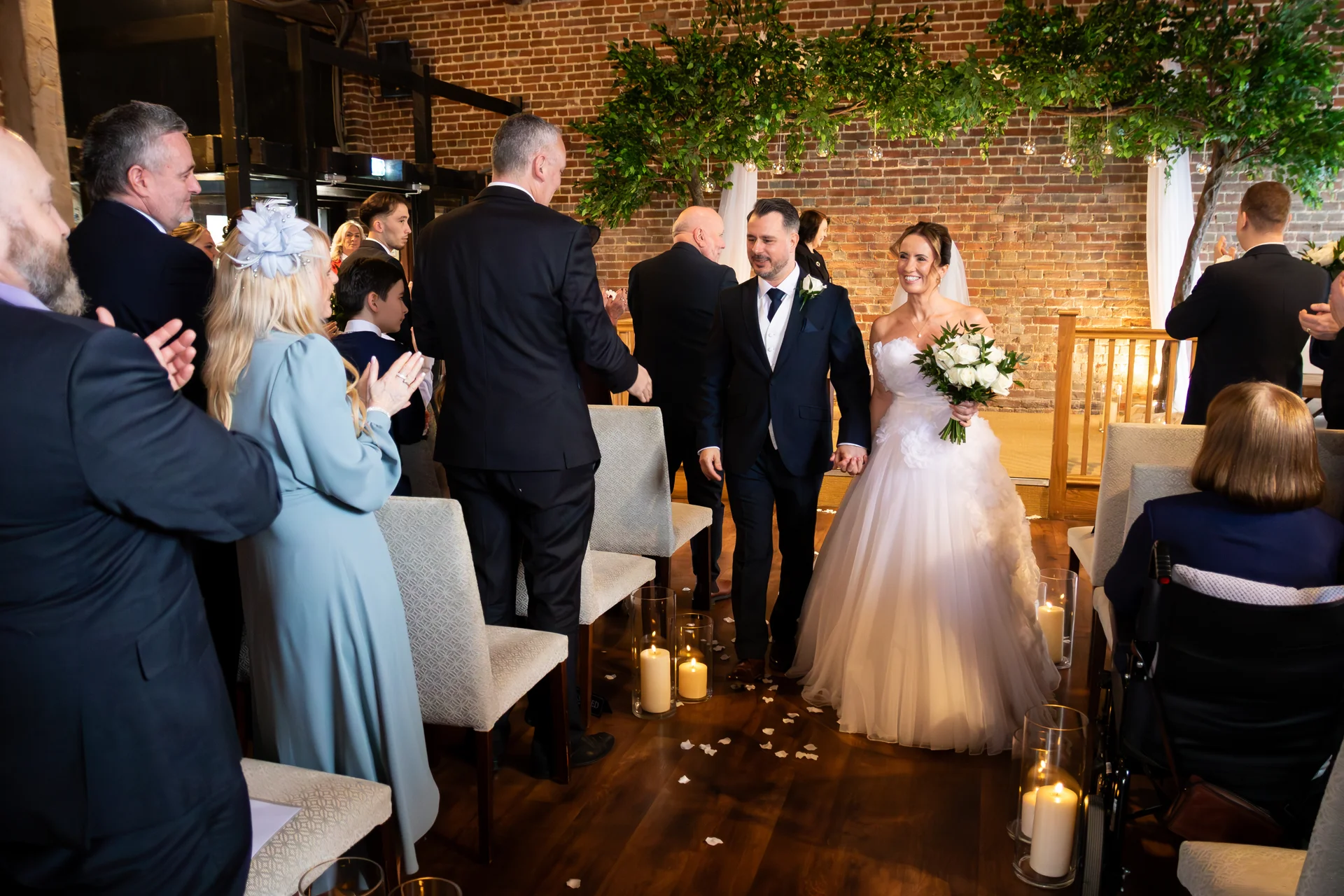 The newly married couple walk hand-in-hand down the aisle together, smiling as cheering guests applaud them in a rustic brick venue decorated with candlelight.