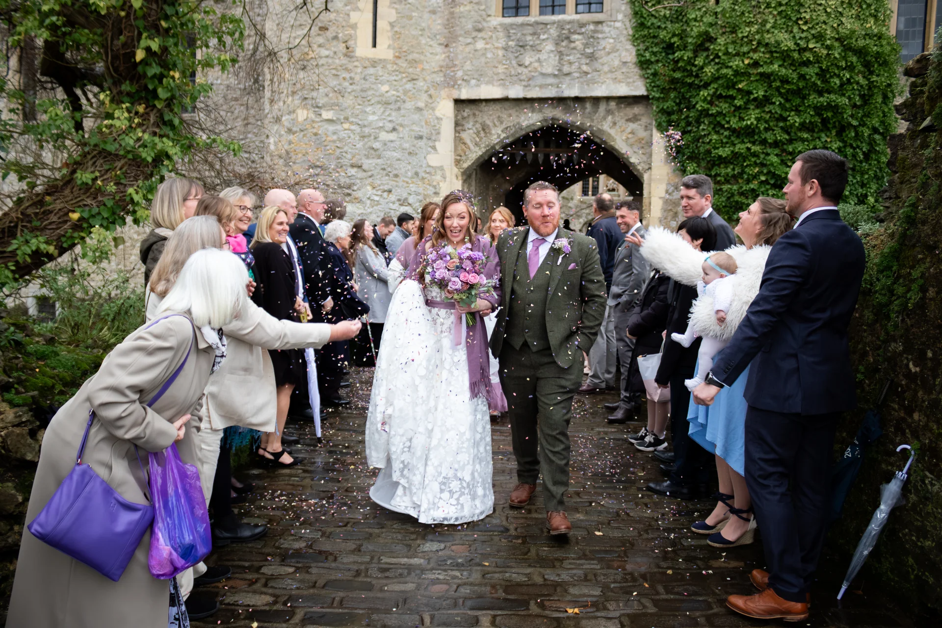 A joyful newlywed couple walks through a historic stone courtyard amidst a shower of colorful confetti, surrounded by cheering wedding guests.