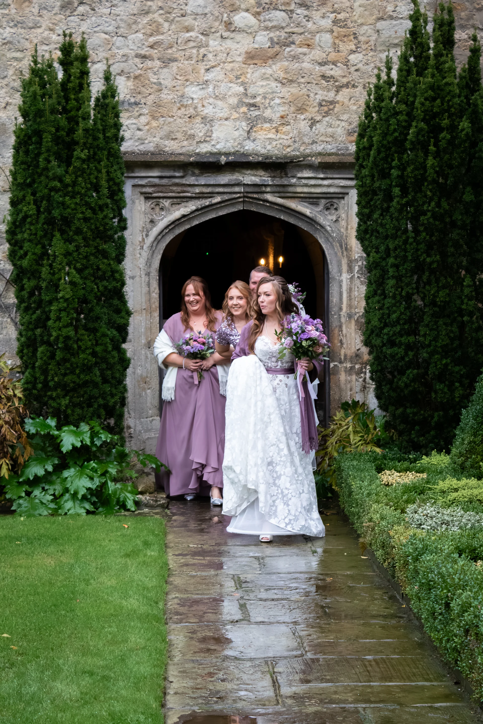 A smiling bride holding a bouquet and lifting her patterned lace dress leads her wedding party exiting through a stone archway flanked by tall trees.