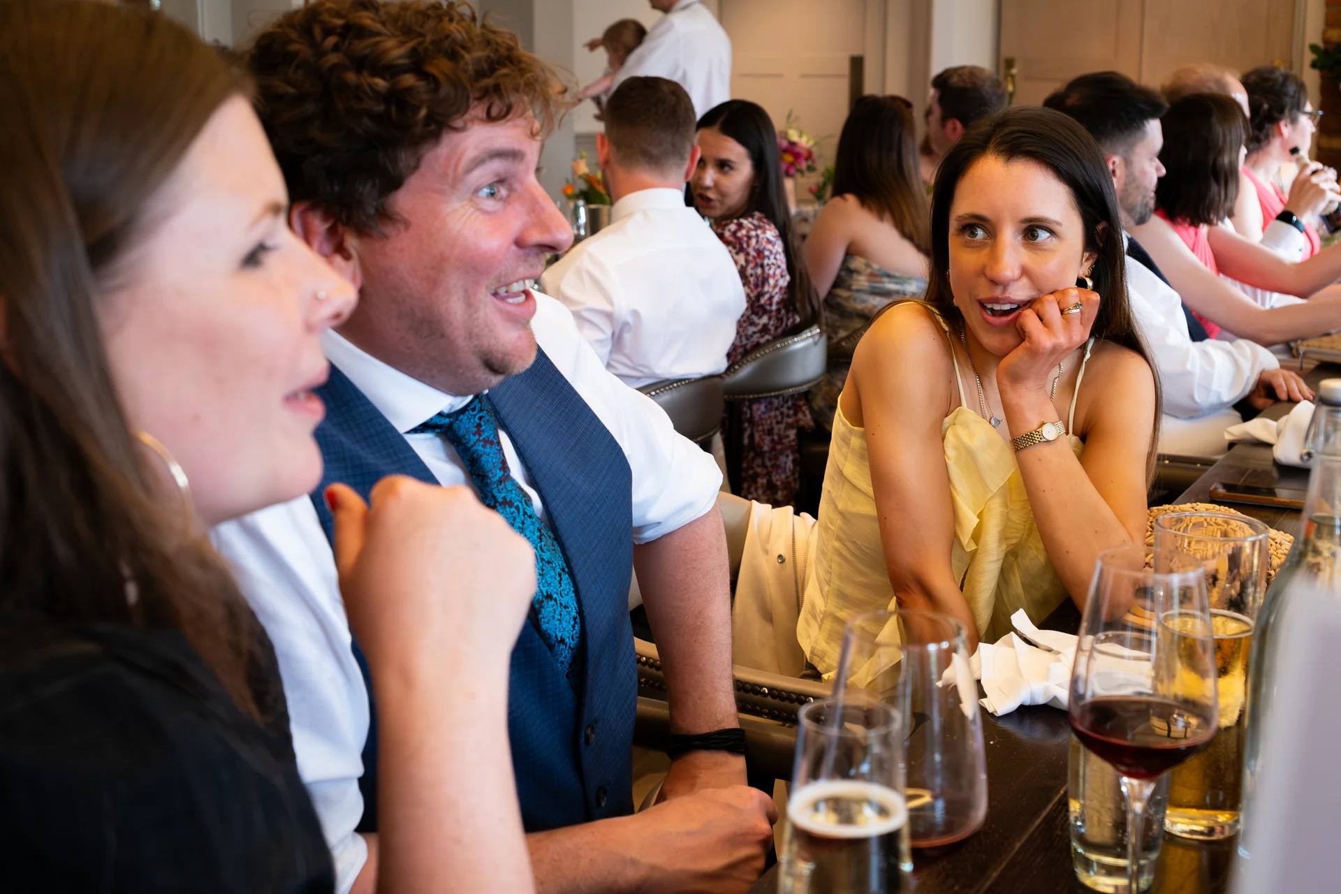 Wedding guests sit at a banquet table during speeches, reacting with amusement and laughter while enjoying drinks.