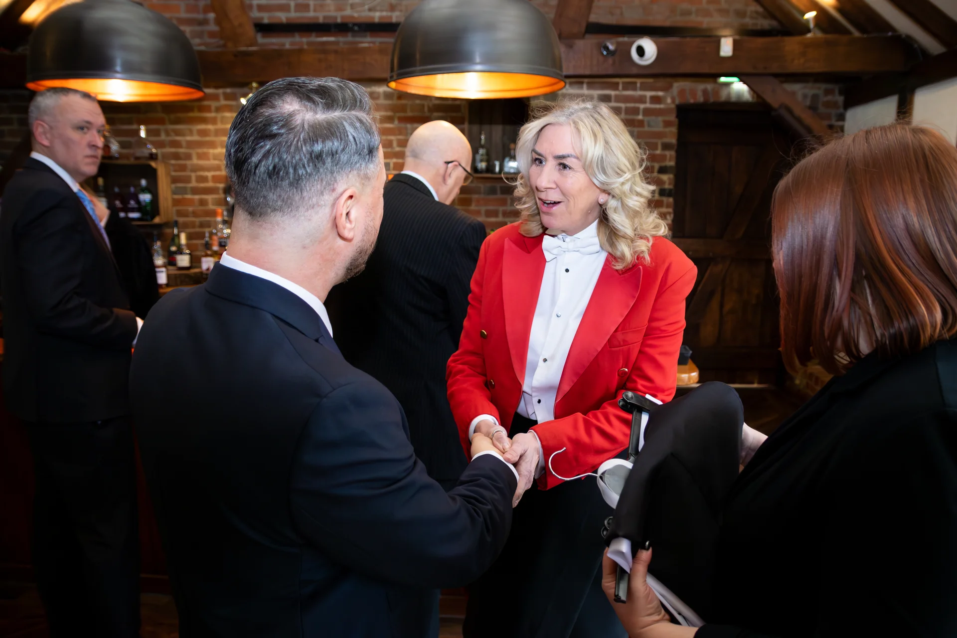 A guest in a dark suit shakes hands with a woman wearing a red jacket and white bow tie inside a rustic venue with exposed brick walls.
