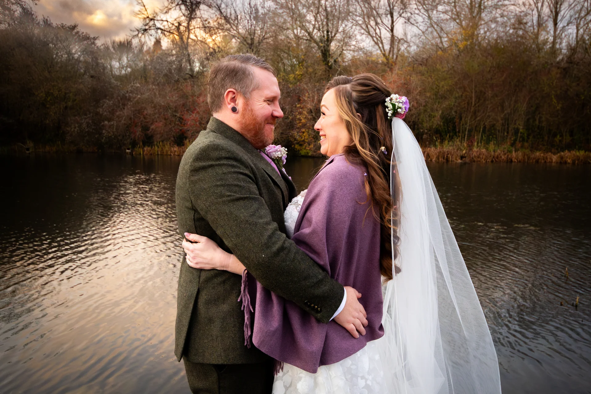 A bride and groom lovingly embrace by a tranquil lakeside during golden hour, with the bride wearing a purple shawl and a veil.