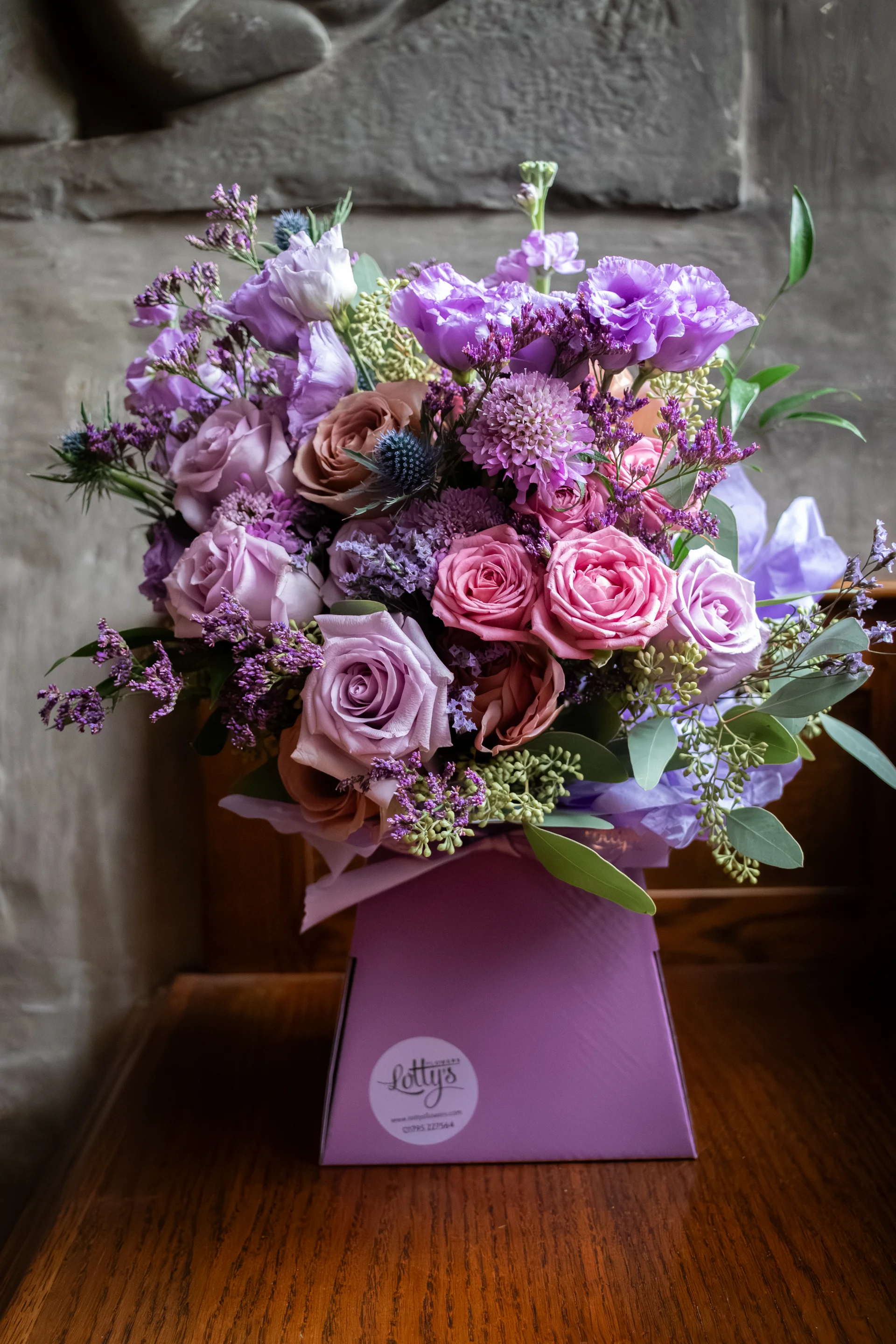An elaborate floral arrangement featuring a mix of purple roses, pink lisianthus, and thistles inside a purple presentation box on a wooden surface.