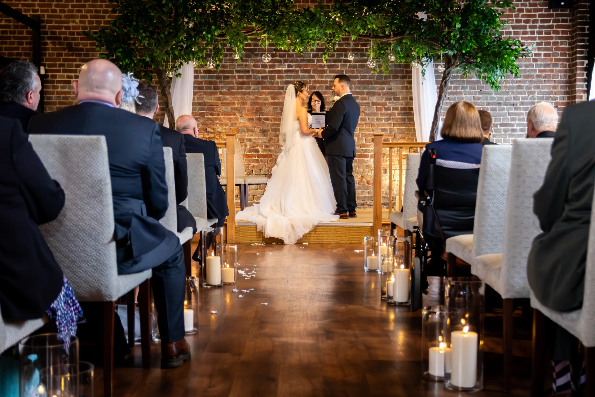 A bride and groom exchange vows during an indoor civil ceremony, framed by an exposed brick wall, overhead foliage installations, and a candle-lined aisle.