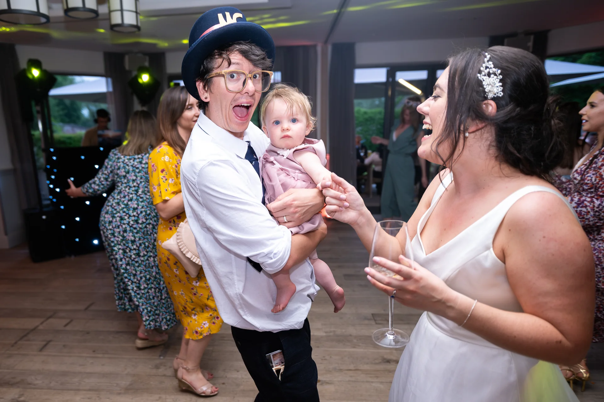 A joyful wedding guest holding a baby while a laughing bride looks on during the evening reception.