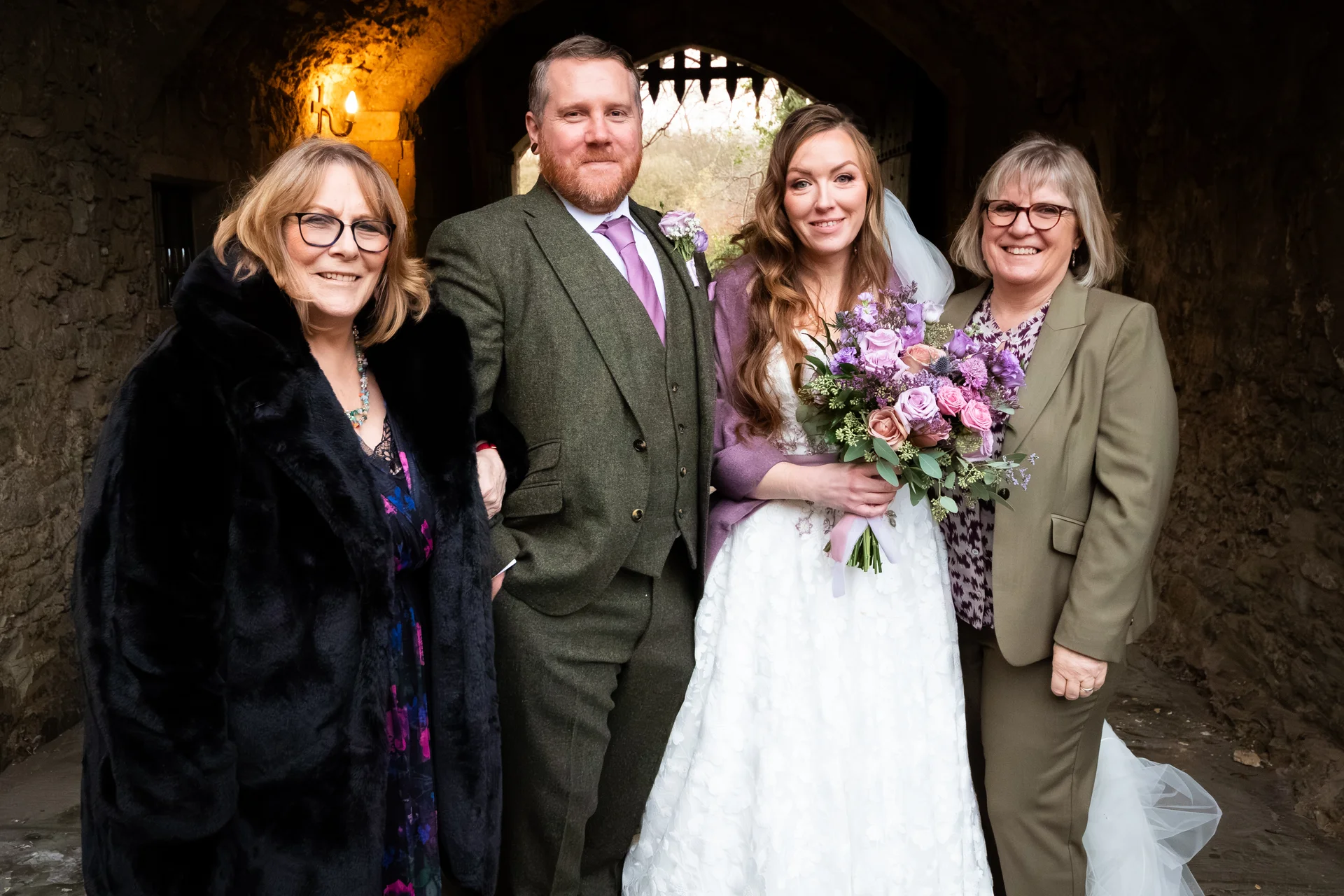 A smiling bride holding a purple floral bouquet stands with her groom in an olive tweed suit and two family members inside a rustic stone castle doorway.