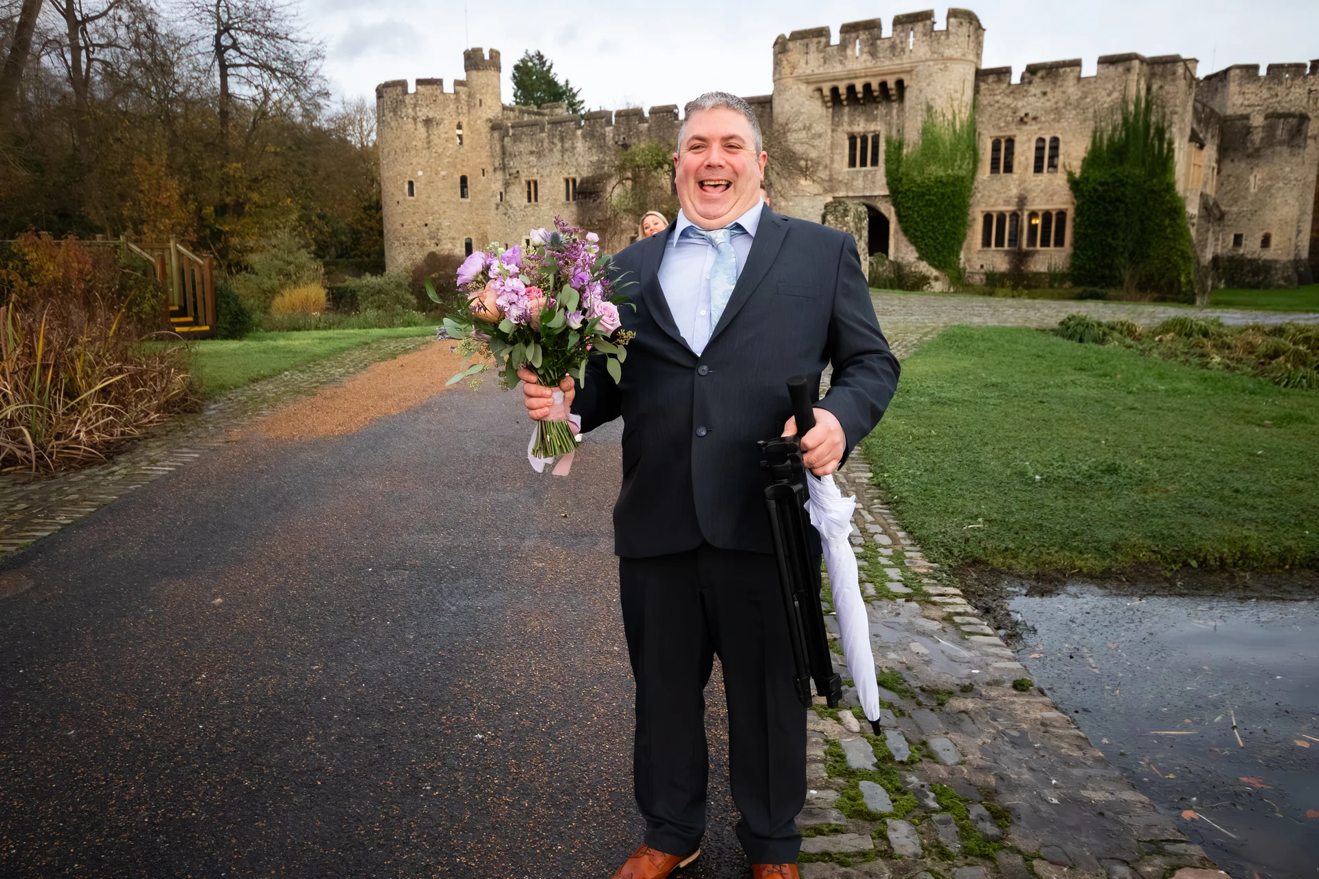 A joyful groom laughs heartily while holding a colourful wedding bouquet and an umbrella in front of a historic stone castle.