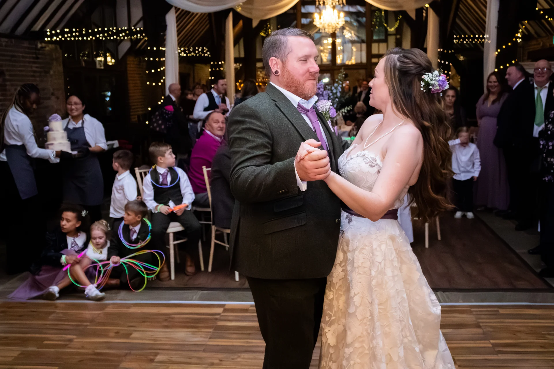 A newlywed couple shares their first dance in a rustic barn venue illuminated by string lights, surrounded by seated guests and children playing with glow sticks.