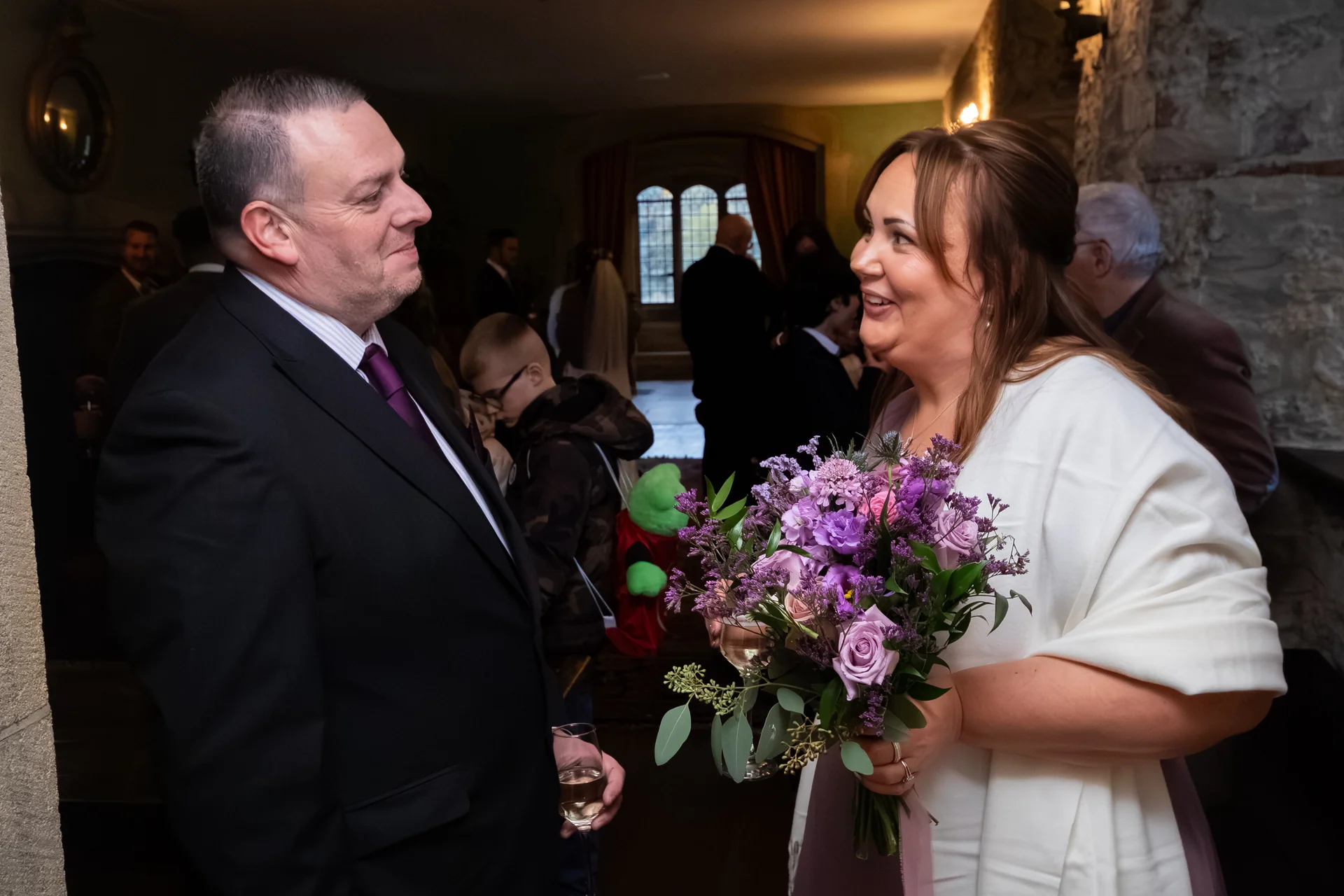 A smiling bride holding a purple floral bouquet and a groom in a black suit look lovingly at each other during a castle wedding reception.
