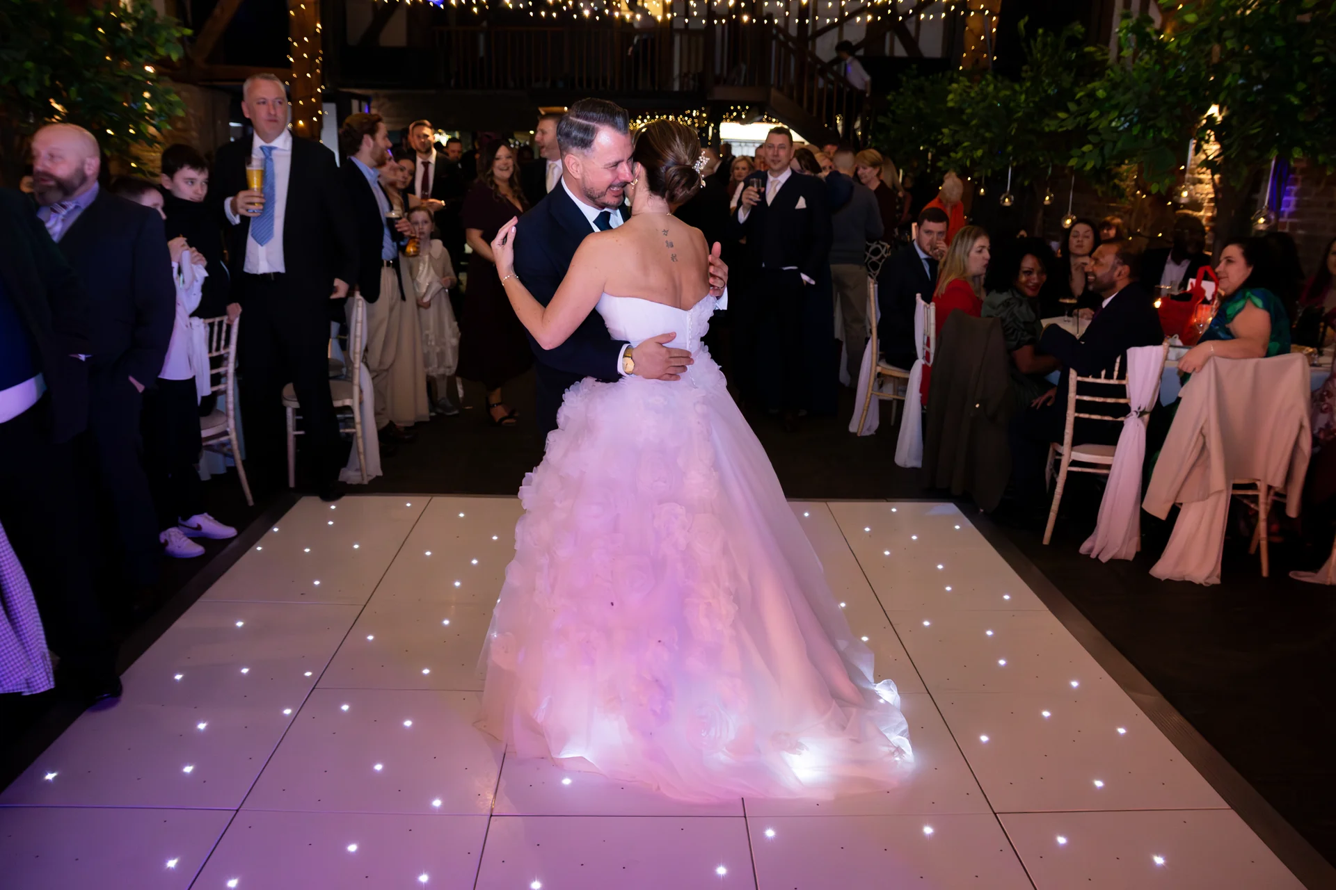 A newlywed couple shares their first dance on an illuminated starlit dance floor inside a rustic barn venue filled with cheering guests.