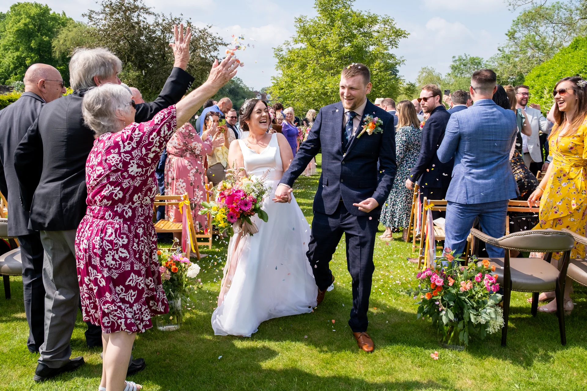 A joyful newlywed couple walks hand in hand through an outdoor garden ceremony as guests cheerfully throw colorful confetti over them.