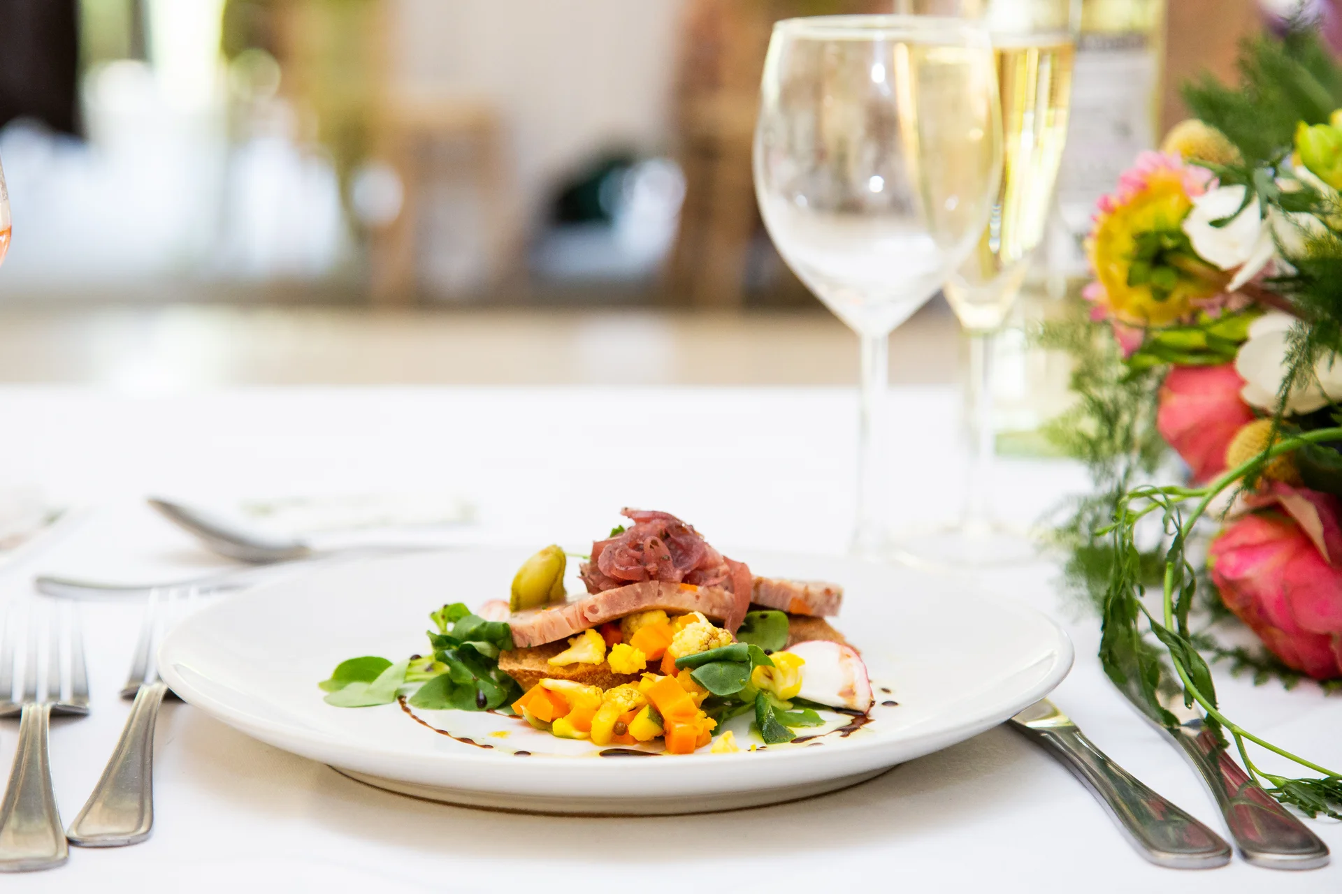 An elegantly plated wedding breakfast starter is served on a white tablecloth, accompanied by wine glasses and a colourful floral centrepiece.