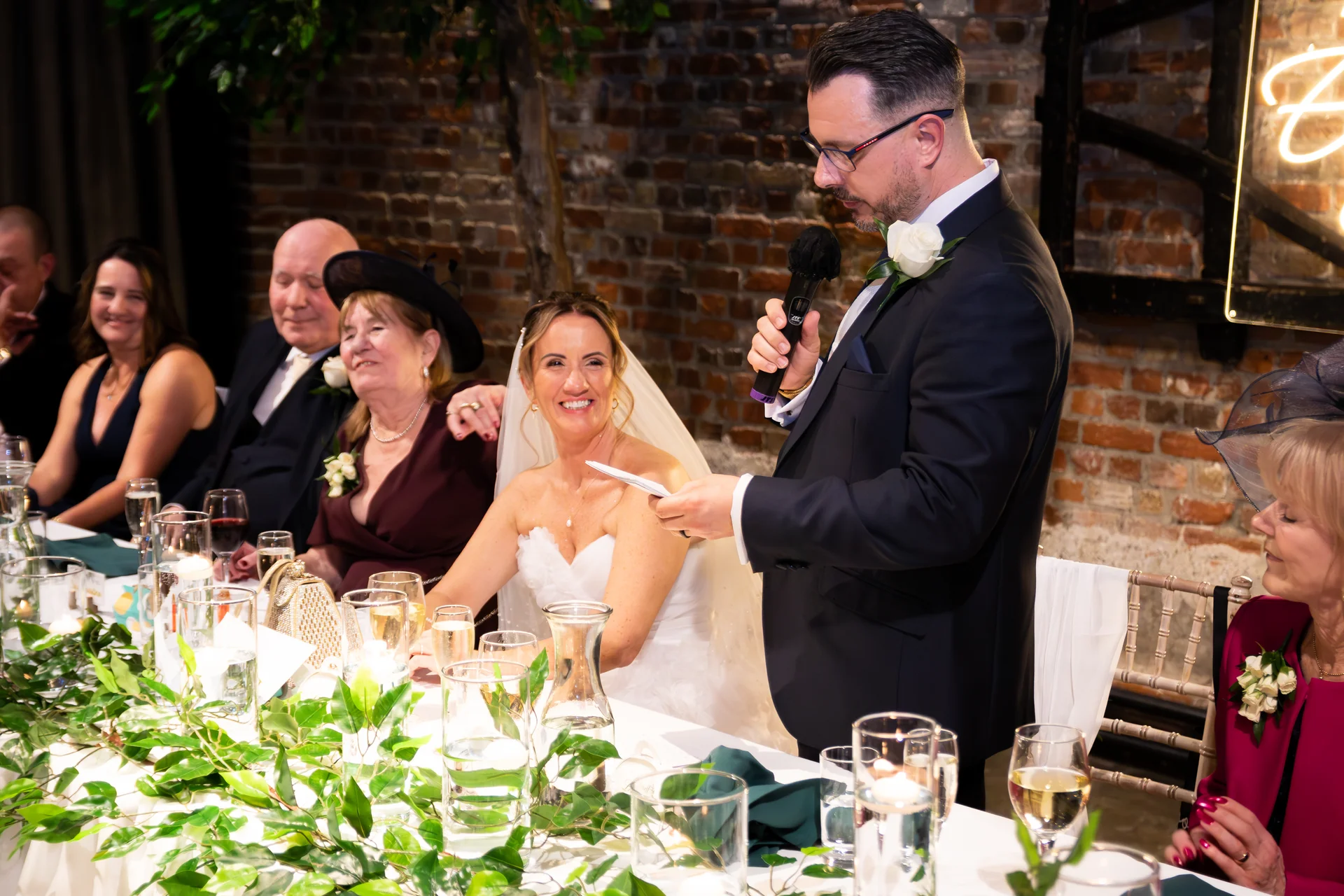 A groom delivers a speech at the top table in a rustic exposed brick venue, while the smiling bride and guests listen closely.