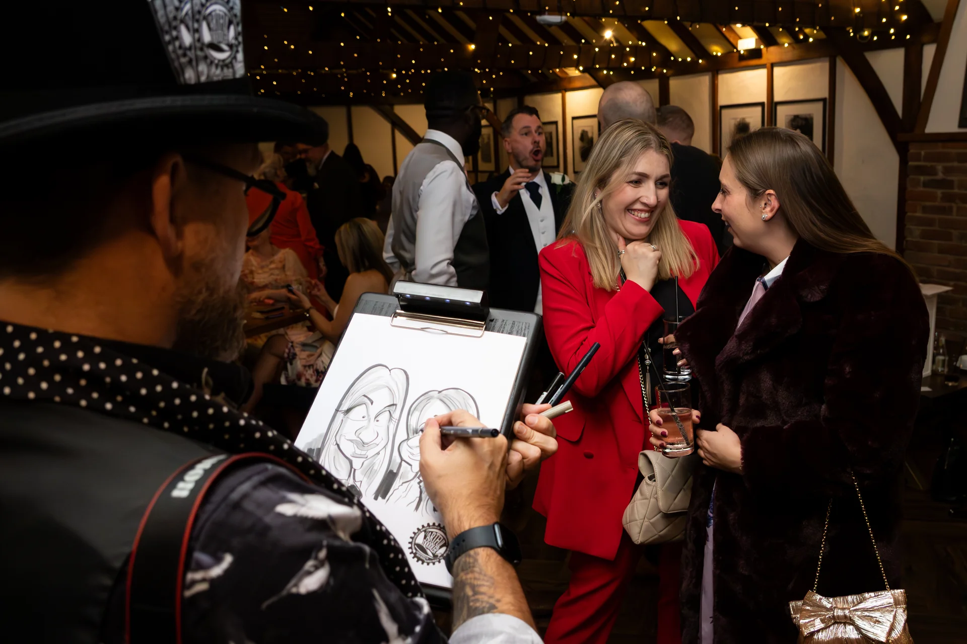 A live caricaturist sketches smiling wedding guests during the evening reception inside a rustic barn decorated with fairy lights.