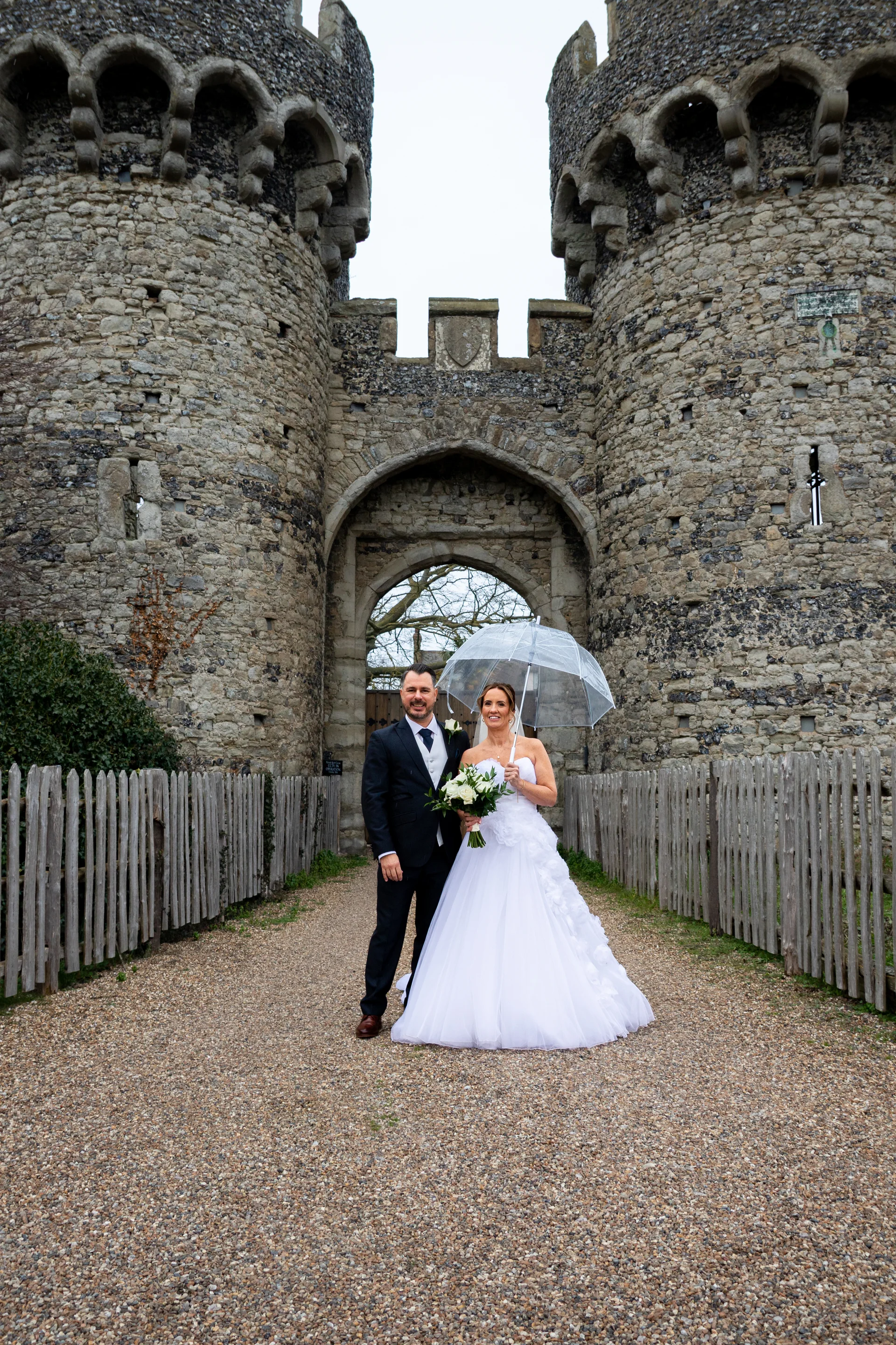 A newlywed couple poses together holding a clear umbrella on a gravel path in front of a grand medieval stone castle gatehouse.