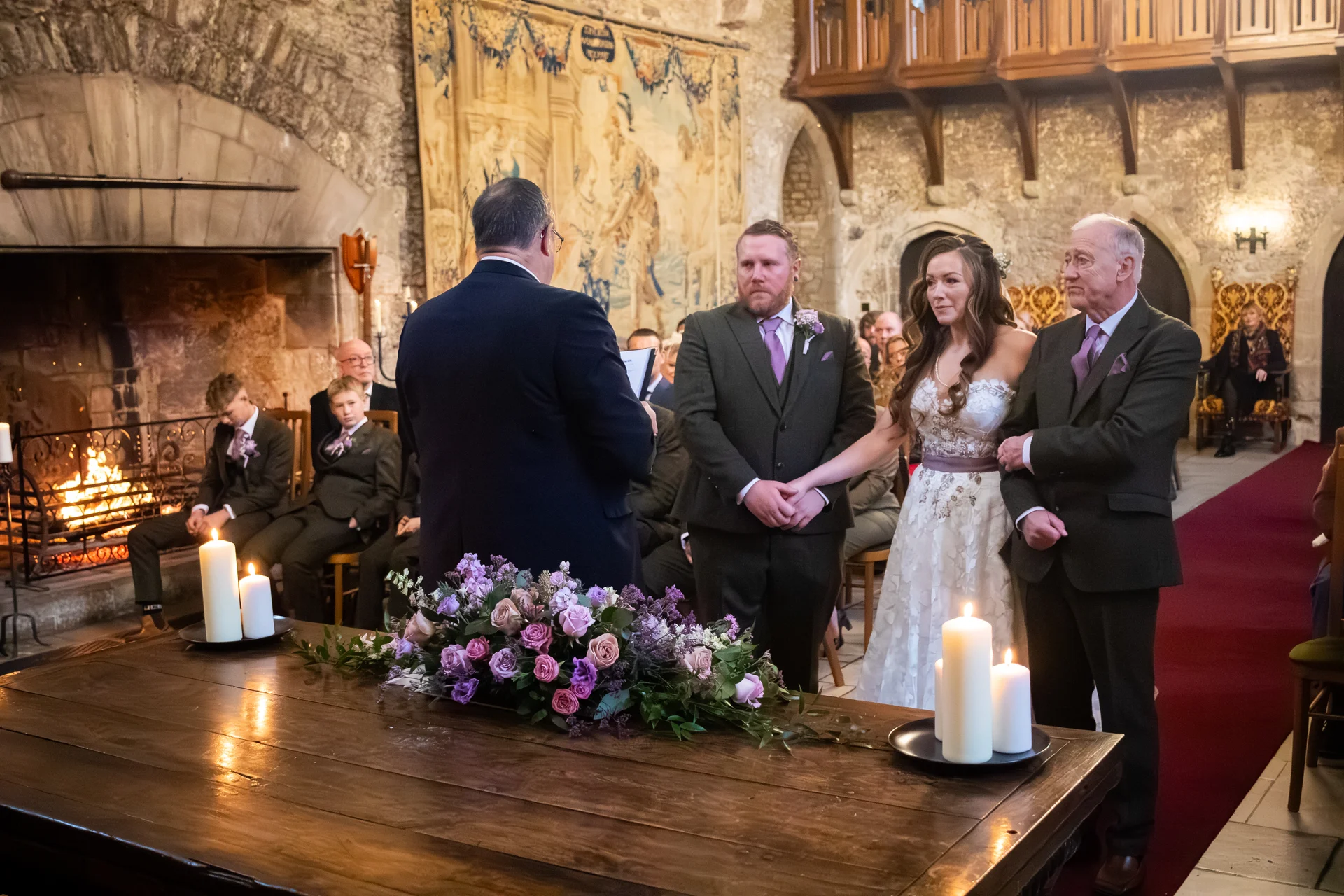 A solemn wedding ceremony takes place inside a historic stone castle hall with a large fireplace, where the bride and groom stand beside a wooden signing table adorned with purple floral arrangements and candles.