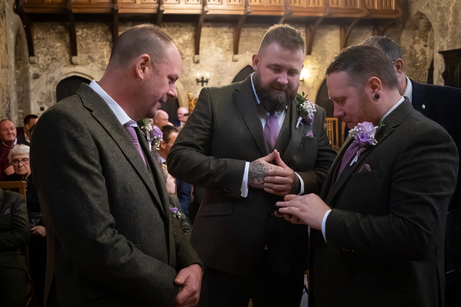 A group of groomsmen wearing matching olive tweed suits and lavender ties share a lighthearted moment inside a historic stone venue.