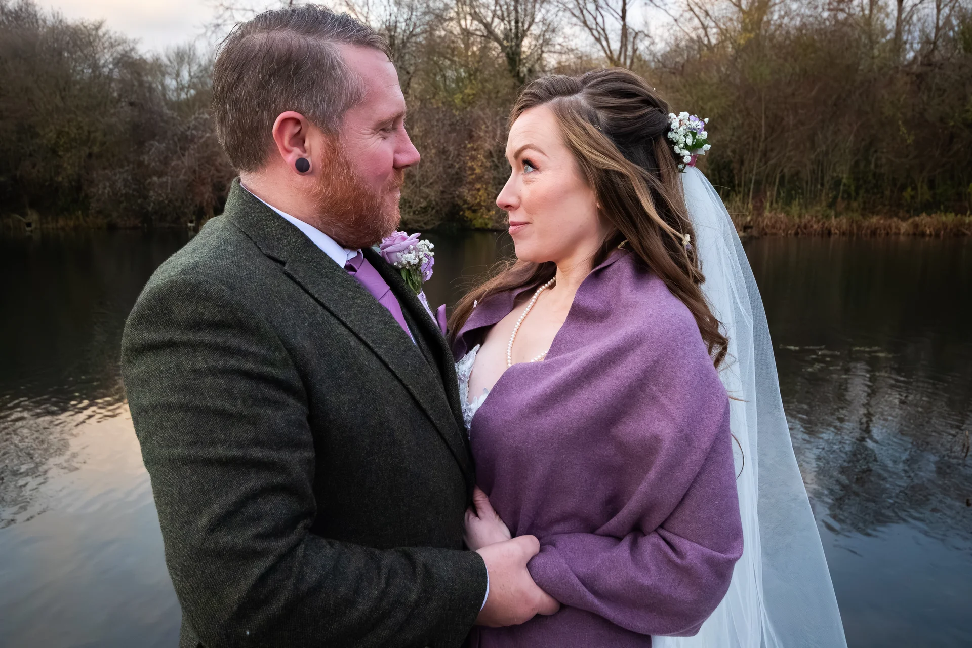 A newlywed couple shares a tender glance by a calm lakeside, with the groom wearing a green tweed suit and the bride wrapped in a purple shawl and veil.
