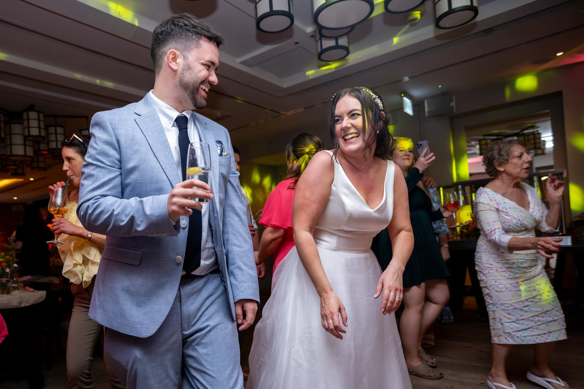 A smiling bride and groom laugh together on a lively dance floor surrounded by wedding guests enjoying the evening reception.