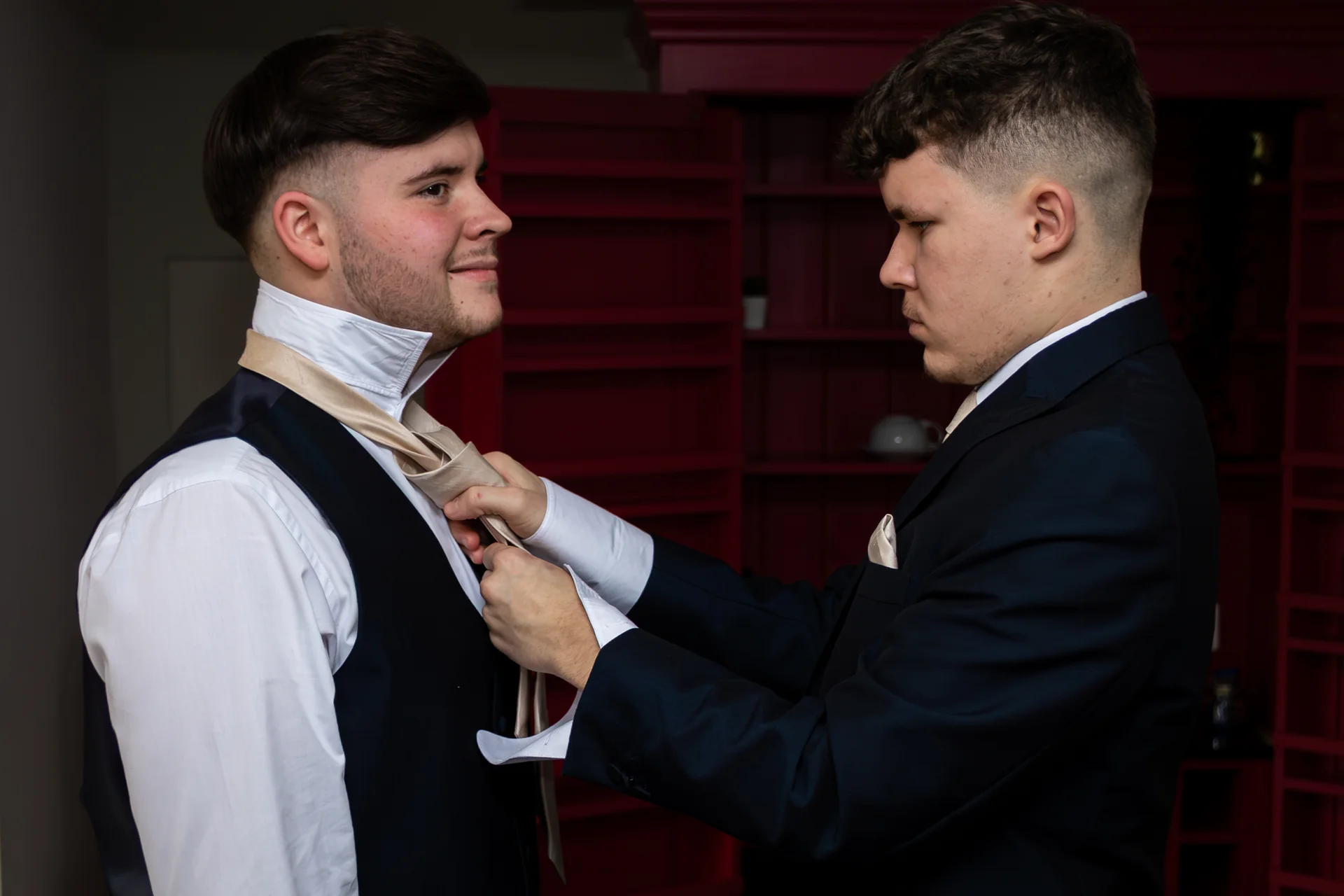 One groom carefully helps adjust and tie his partner's tie during wedding morning preparations indoors.