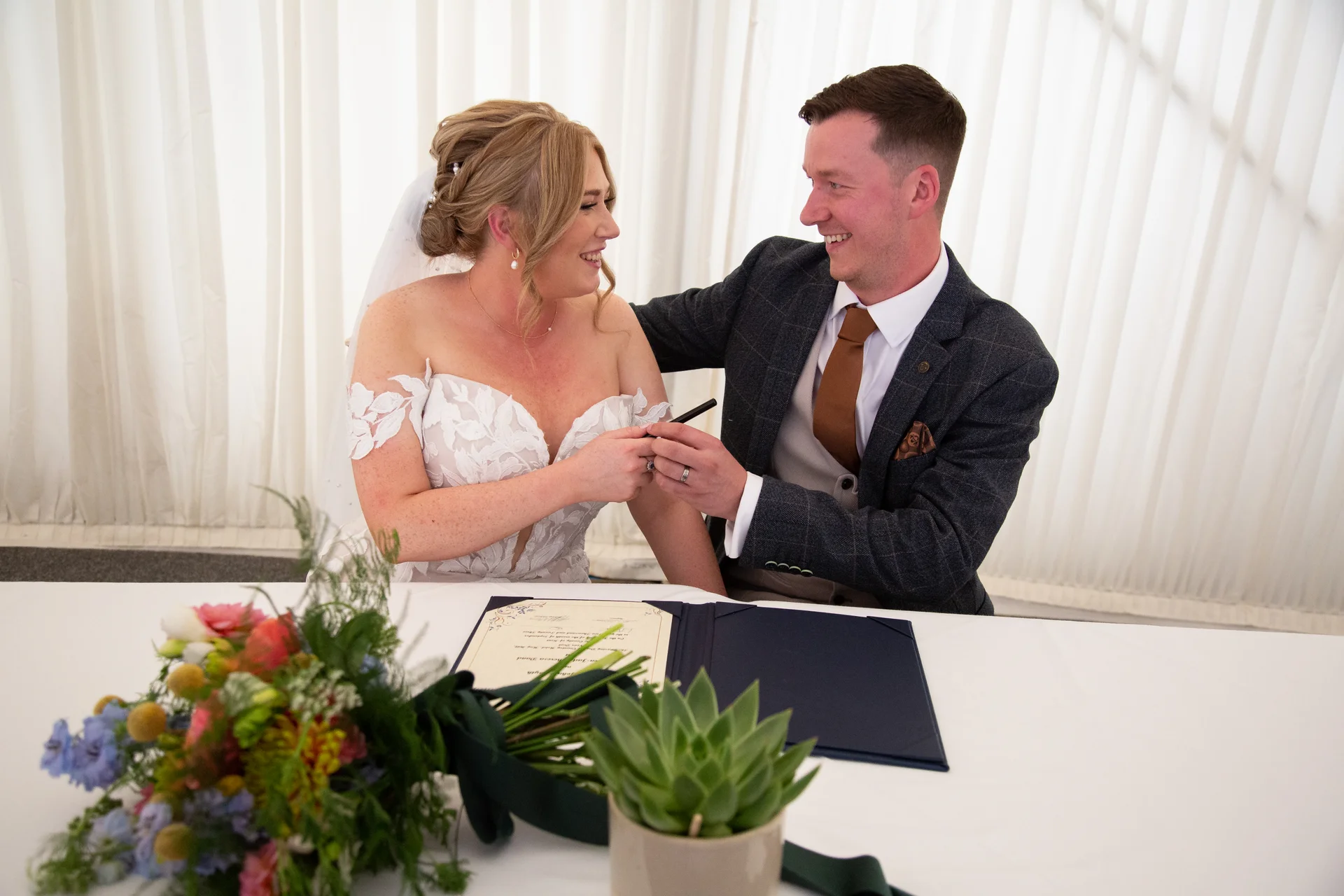 The bride and groom smile happily at each other while signing the marriage certificate during their wedding ceremony inside a marquee.