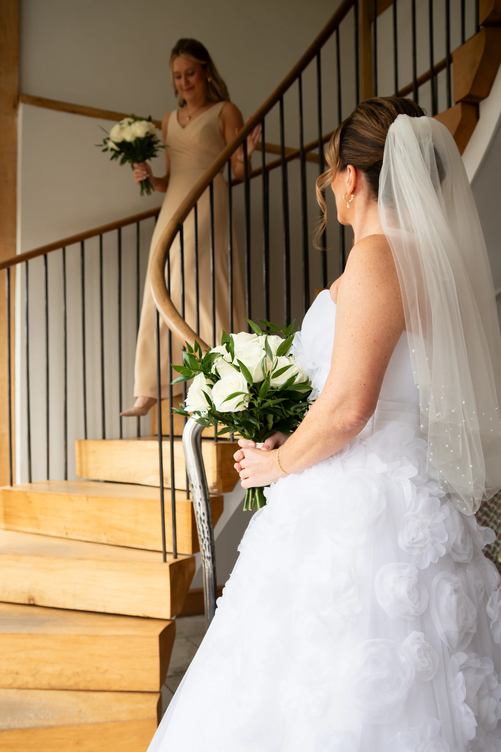 A bride in a white strapless gown with floral appliques and a veil holding a rose bouquet while a bridesmaid walks down the wooden spiral staircase behind her.